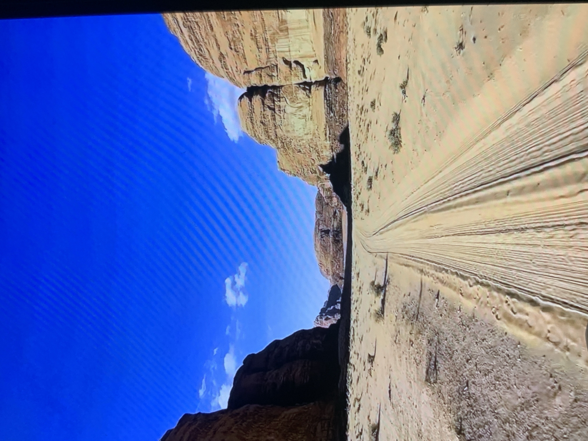 The image depicts a vast, arid landscape with towering sandstone cliffs and a clear blue sky dotted with a few white clouds. A dry, sandy track stretches into the distance, showing visible tire tracks. Sparse, low-lying desert vegetation is scattered across the sandy terrain. The scene conveys a sense of isolation and the harsh beauty of a desert environment. The lighting suggests it is daytime, likely mid-morning or afternoon, given the bright sun and distinct shadows cast by the cliffs. There is no visible text in the image. The orientation of the image is rotated 90 degrees counter-clockwise.