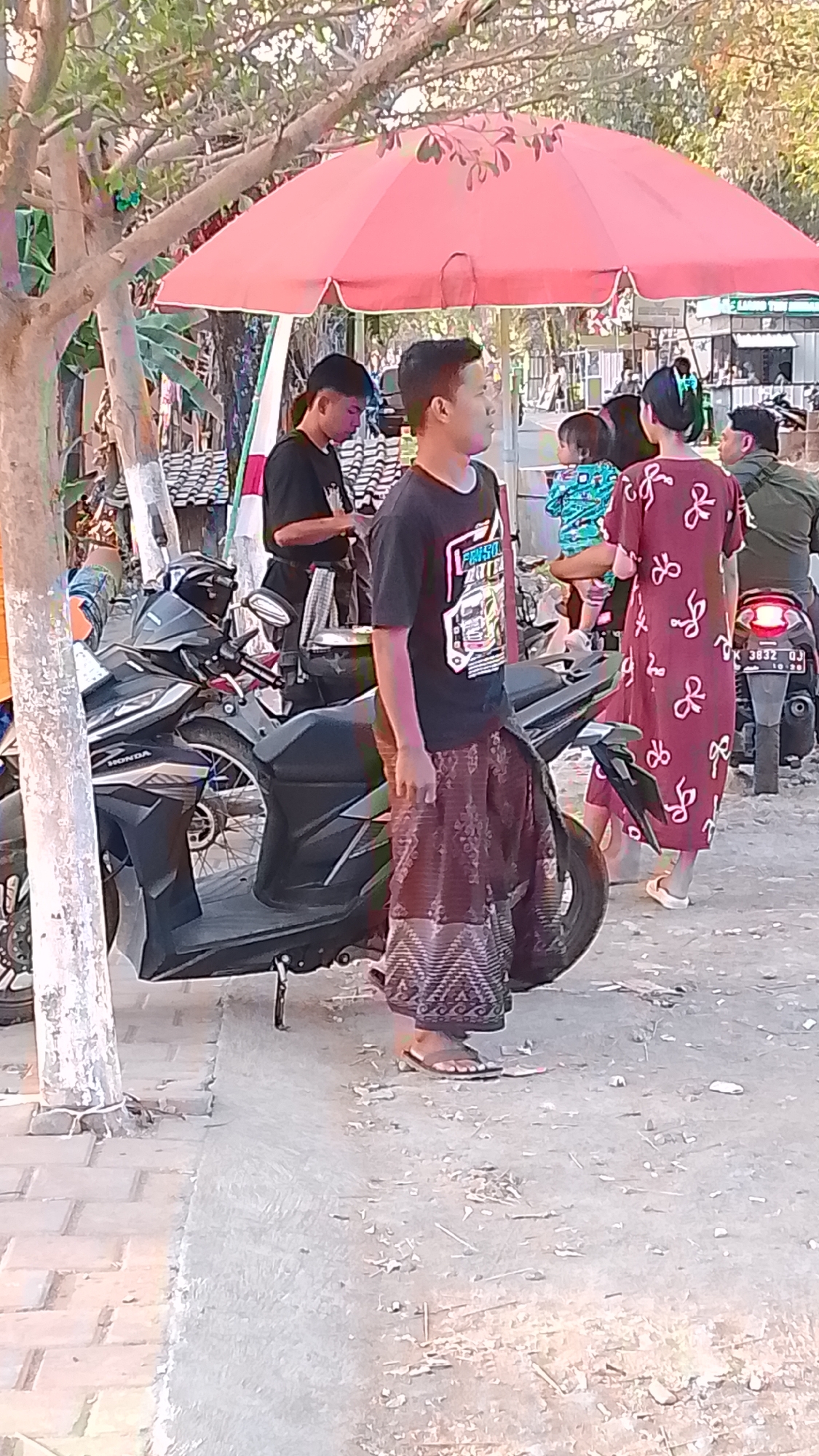In Karanglangu, Indonesia, a group of people are gathered outdoors near a street. A dark grey Honda scooter is parked on the left side of the frame. A young man in a black t-shirt with a graphic print and patterned pants stands next to the scooter, facing right. To his left, another young man in a black t-shirt is visible. Behind them, a woman in a maroon dress with a white bow pattern and a child in a blue shirt are standing near another scooter. Further back, another individual wearing a green shirt is seated on a motorcycle. A large red umbrella provides shade over part of the scene, and trees with green foliage are visible in the background. The ground is a mix of paved and dirt surfaces. The overall scene suggests a casual gathering or stop.