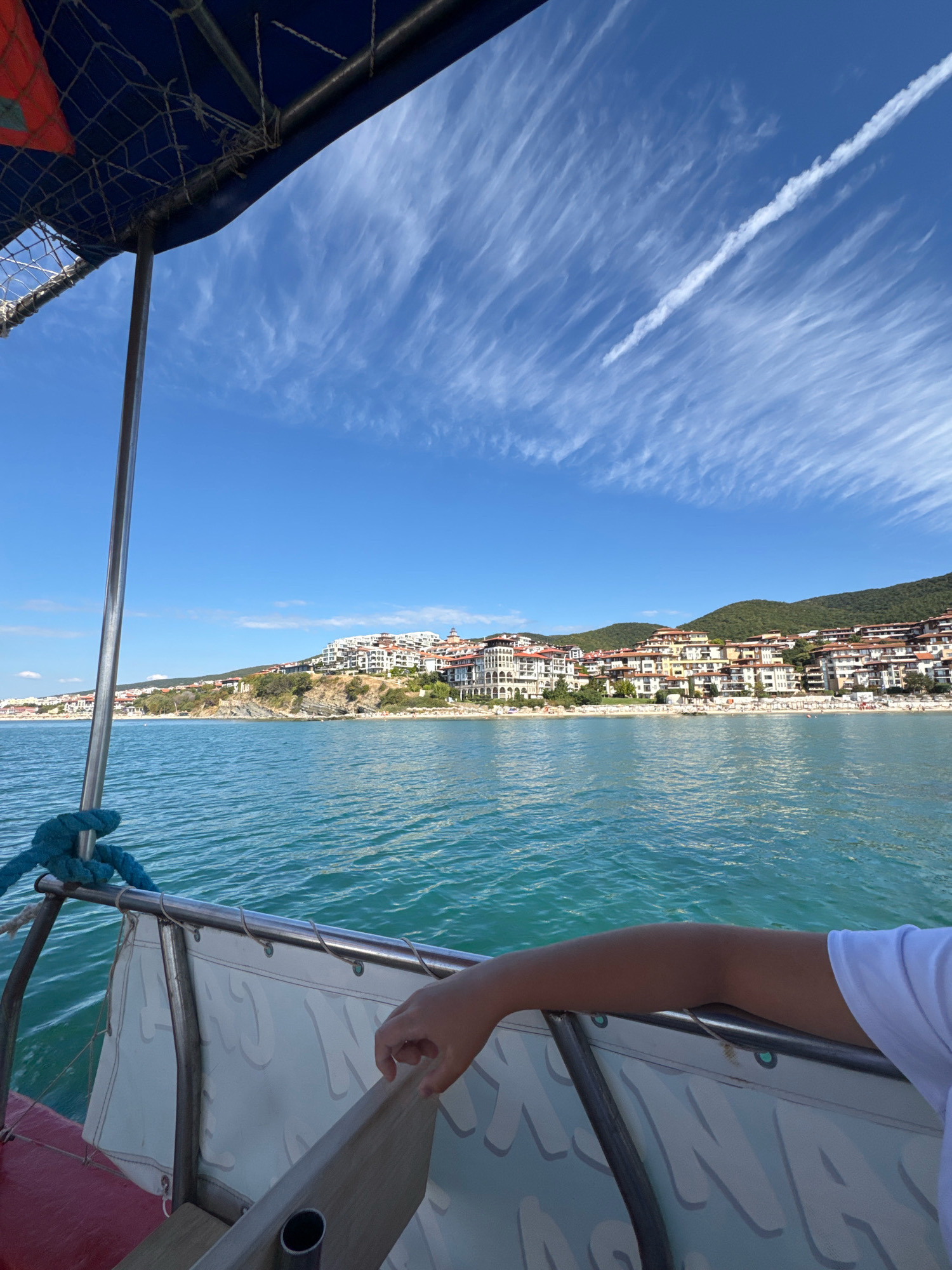 The image depicts a view from a boat sailing on the Black Sea towards the city of Sveti Vlas, Bulgaria. The city is situated on a hillside overlooking the sea, with numerous white buildings and red-tiled roofs cascading down towards a sandy beach. The water is a vibrant turquoise, reflecting the clear blue sky dotted with wispy white clouds. A jet of condensation from an aircraft streaks across the sky. In the foreground, the edge of the boat is visible, with a blue rope tied to a metal railing. A person's arm, wearing a white t-shirt, rests on the railing, with their hand gently touching a wooden plank. The boat appears to be a small passenger vessel, possibly used for tours. The overall scene conveys a sense of leisure and scenic beauty.