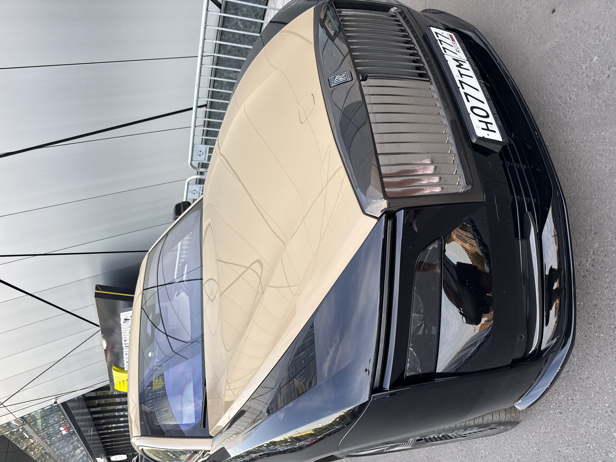 A black and beige Rolls-Royce Phantom, distinguished by its dual-tone paint job, is parked on a paved street in Moscow, Russia. The car's front grille, hood, and headlights are prominently visible. A license plate with the identifier "H077TM 777 RUS" is affixed to the front bumper. Behind the car, a modern building with a metallic facade and glass windows creates a backdrop, while a metal fence stands between the car and the building. The scene is illuminated by daylight, casting reflections on the car's polished surfaces.