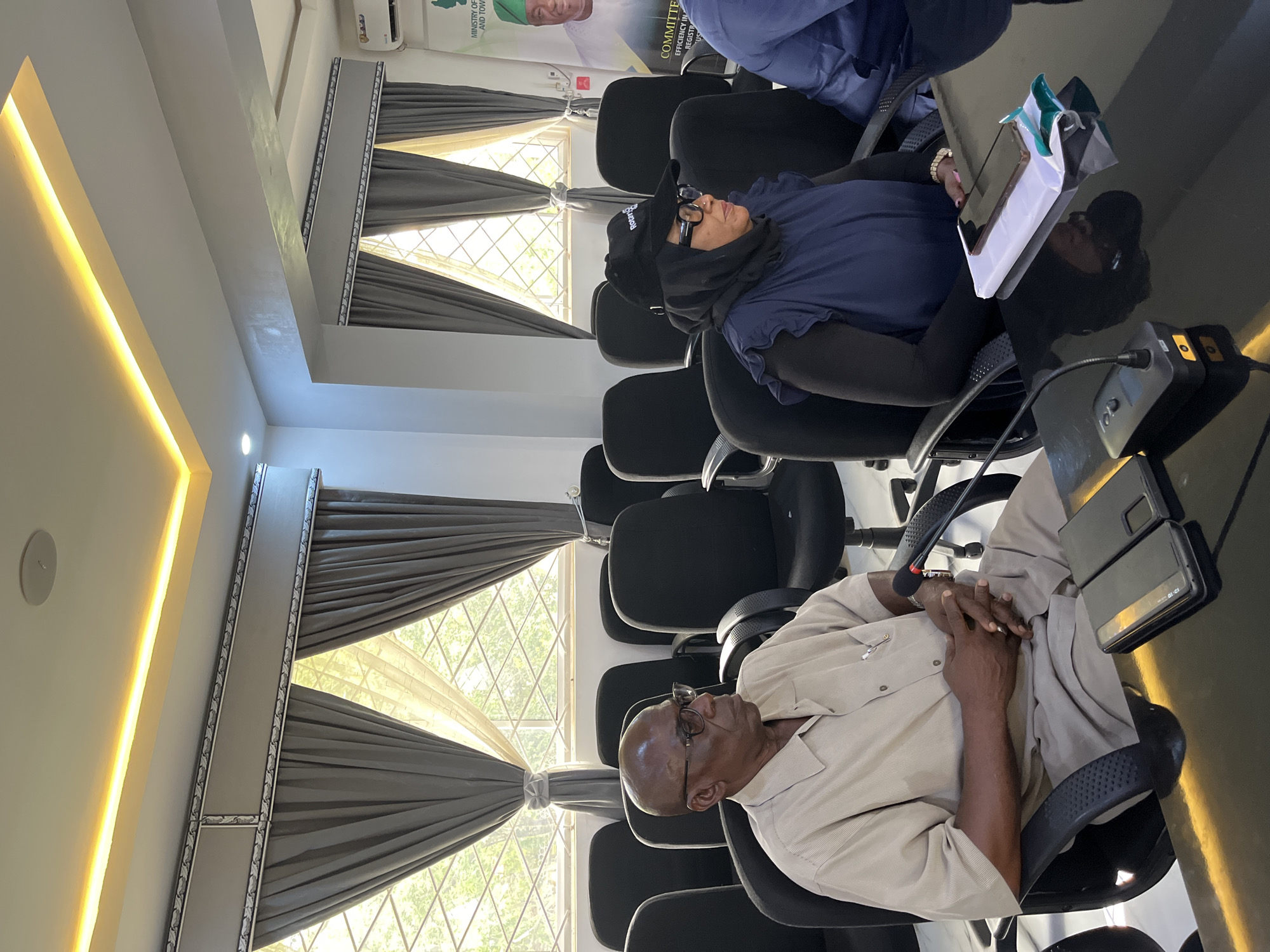 Two individuals are seated in a conference room, facing the camera from the chest up. On the left, an older Black man wearing glasses and a light brown collared shirt with a pen clipped to the pocket is looking to his right. He has his hands clasped in his lap. On the right, a Black woman wearing a dark blue vest over a black long-sleeved shirt, a black headscarf, and a black baseball cap with a white logo is looking to her right. Her hands are clasped in her lap. In front of them are two black microphones, a black table, and a tablet. The room has gray curtains covering diamond-patterned windows and an illuminated ceiling. A poster is visible on the right wall featuring a man in traditional attire and text that reads "MINISTRY OF FINANCE AND TOWN PLANNING". This image was captured at the Plateau State Ministry of Finance and Economic Planning conference room in Jos, Nigeria.