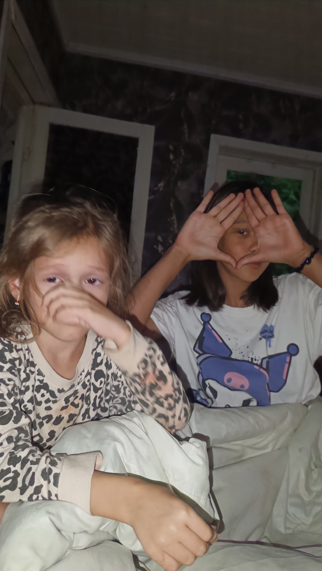 Two young females are seated on a light-colored bed. The female on the left, with light brown hair, is wearing a long-sleeved shirt with a leopard print. Her right hand is touching her face, obscuring her mouth. The female on the right, with dark brown hair, is wearing a white t-shirt with a blue cartoon character graphic on it. She is holding her hands up in front of her face, forming a triangular shape. The background appears to be a room with dark, textured walls and two framed windows. One window on the left has a white frame and is partially open, revealing a darker interior. The window on the right is also framed in white. The lighting is dim, suggesting it may be nighttime or an indoor setting with low light. There are no discernable elements indicating the location is in the city of Karsha, Russia.