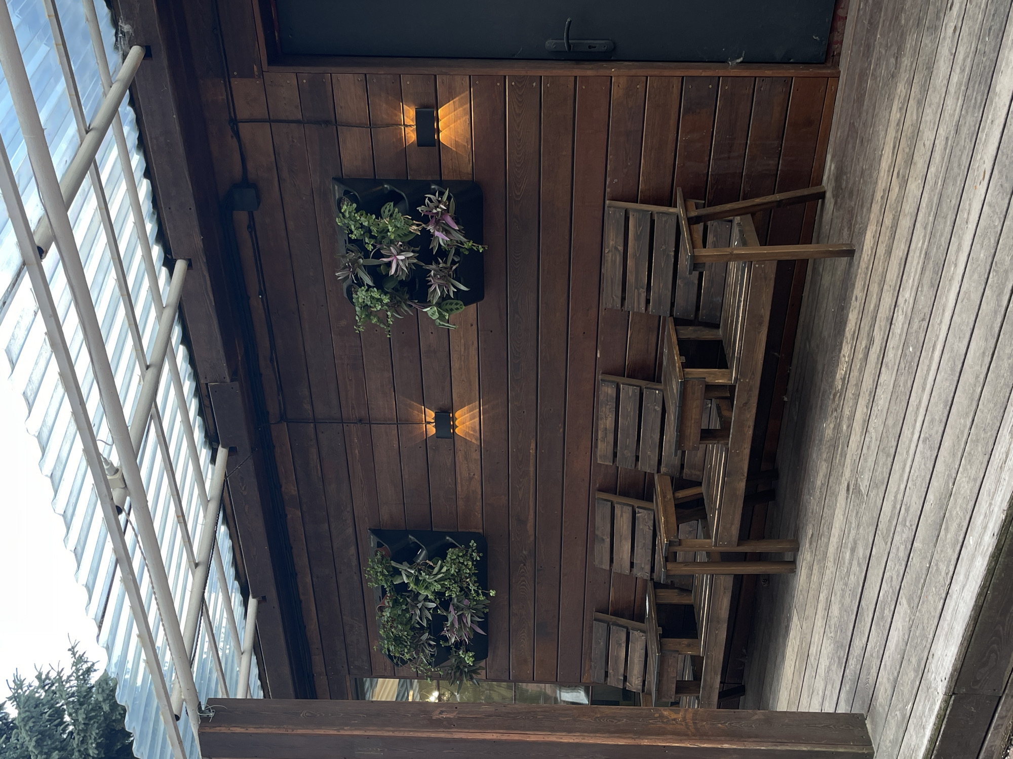 The image depicts an outdoor seating area with wooden benches against a dark wooden wall. Above the benches, two square vertical planters filled with greenery are mounted on the wall. Between the planters, two modern outdoor wall lights are installed, casting warm light. A corrugated translucent roof is visible overhead, supported by metal beams. A dark grey door is on the right side of the wall. The wooden deck in the foreground shows signs of wear. This scene is likely a part of a cafe or restaurant in Moscow, Russia, designed to offer a relaxed outdoor experience.