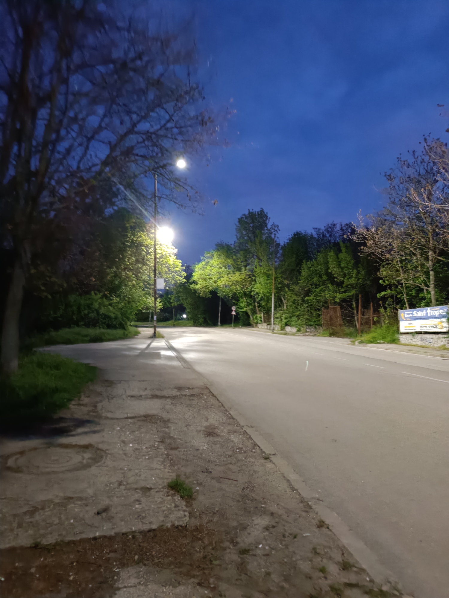 The image depicts an empty road and sidewalk at night or during deep twilight, illuminated by streetlights. A paved road stretches into the distance, gently curving to the right, with a concrete sidewalk and grassy verge on its left. Numerous trees and bushes line both sides of the road; some foliage appears brightly lit and vibrant green from the streetlights, while other trees, particularly on the upper left and right, show bare branches against the dark blue sky.
A prominent streetlight stands on the left side of the road, casting long shadows onto the sidewalk and road surface. The light creates bright reflections on the wet-looking pavement directly beneath it. On the right side of the road, further down, a large billboard is visible behind some trees and a fence. The billboard features text, with "Saint Tropez" clearly visible at the top, and "AUTO AFRICA" or similar text below it.
The sky is a deep, clear blue, characteristic of night or deep dusk. The scene is devoid of people or vehicles, conveying a sense of stillness and quiet. Notable details include a manhole cover on the left-hand sidewalk and the stark contrast between the bright artificial light and the surrounding darkness.