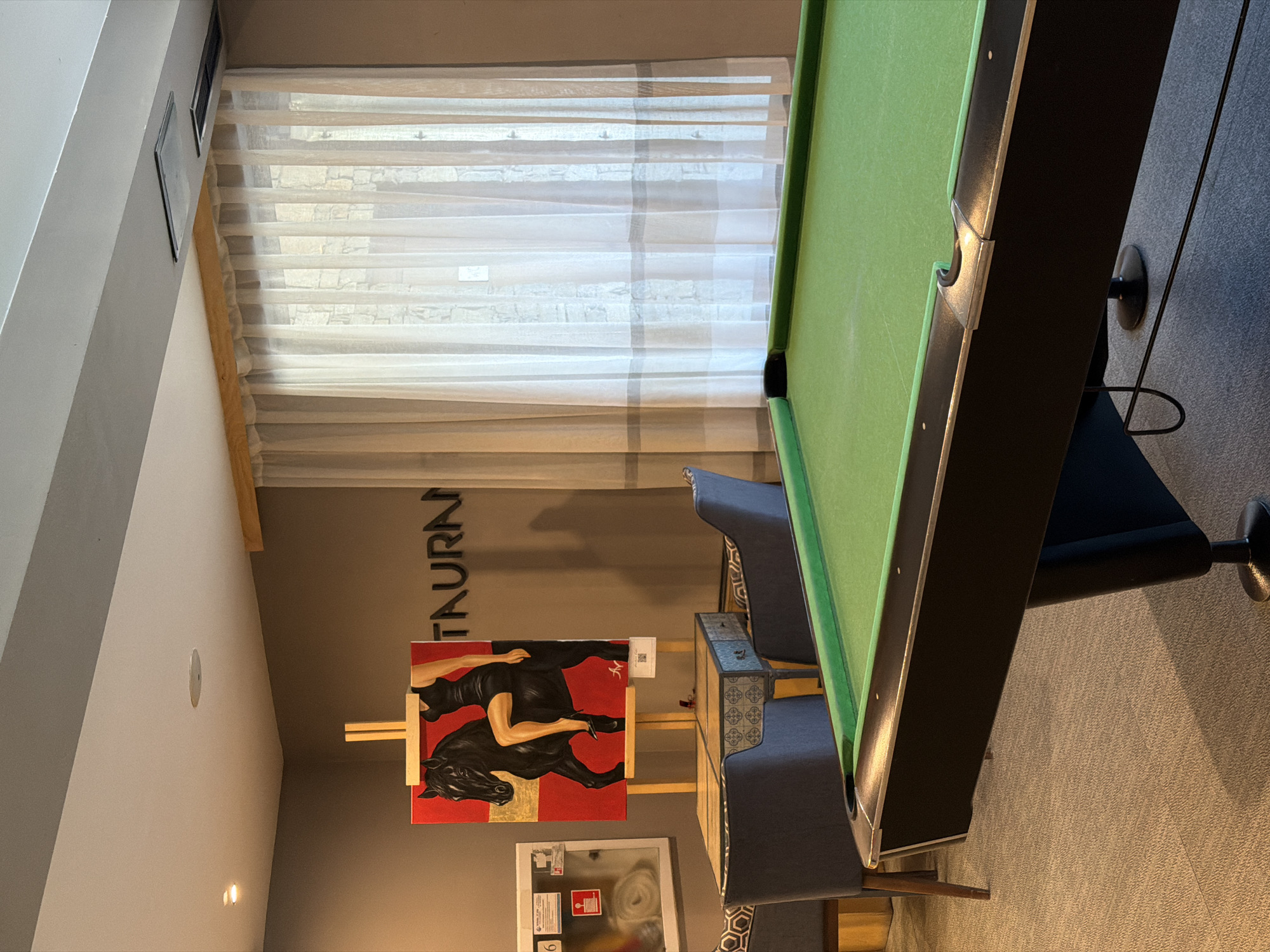 The image shows an interior space with a pool table as the dominant feature. The pool table has a green surface and dark wooden rails. Adjacent to the pool table is a cushioned seating area with blue-gray upholstery. A painting on an easel is also visible, featuring a black horse and human figure against a red and gold background. The wall behind the painting has lettering that partially reads "TAURN". A window with sheer curtains provides natural light. Further details include framed pictures on the wall, and a sign on the wall below the painting. The location is Unknown, Unknown.