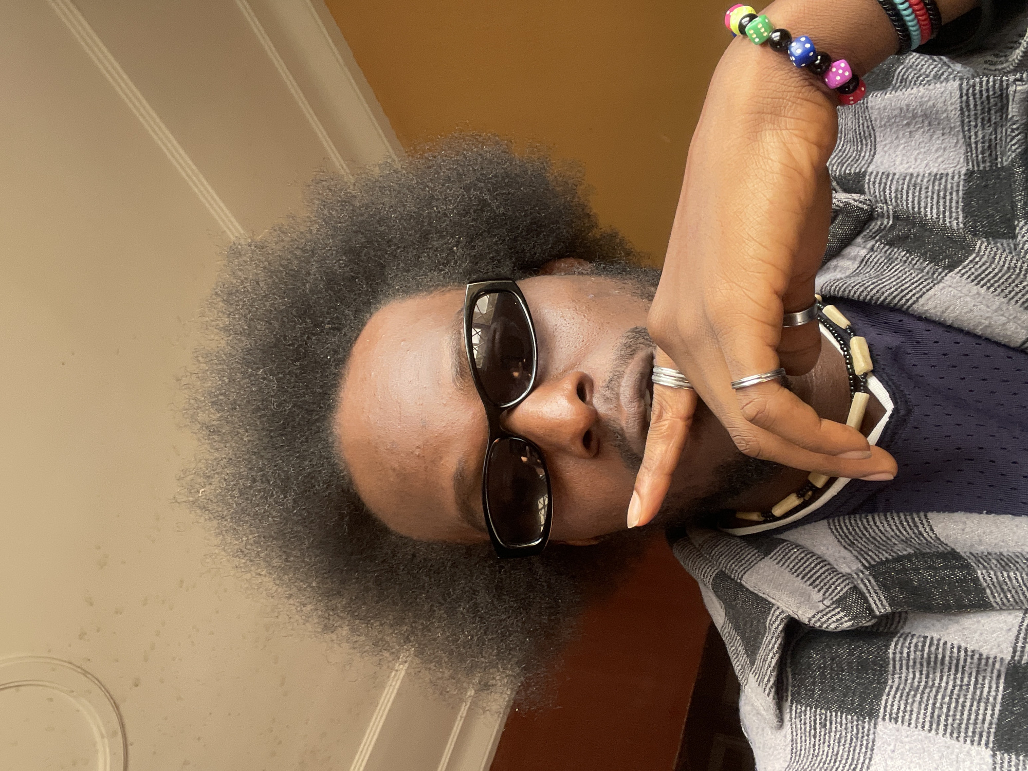 The image is a close-up selfie of a Black man with an afro hairstyle and sunglasses. He is wearing a grey and black plaid shirt over a dark blue mesh-fabric shirt. He has a necklace made of wooden beads and another beaded necklace. He is also wearing several rings on his fingers and a colorful bracelet made of dice-shaped beads on his wrist. His right index finger is touching his lips. The background is a light brown wall with some subtle texture and a white ceiling with molding. The lighting suggests an indoor setting, possibly a room with natural light coming from the side. The image is sharp and in focus, with a shallow depth of field that blurs the background. There is no information about the location in Jos, Nigeria, in the image itself.
