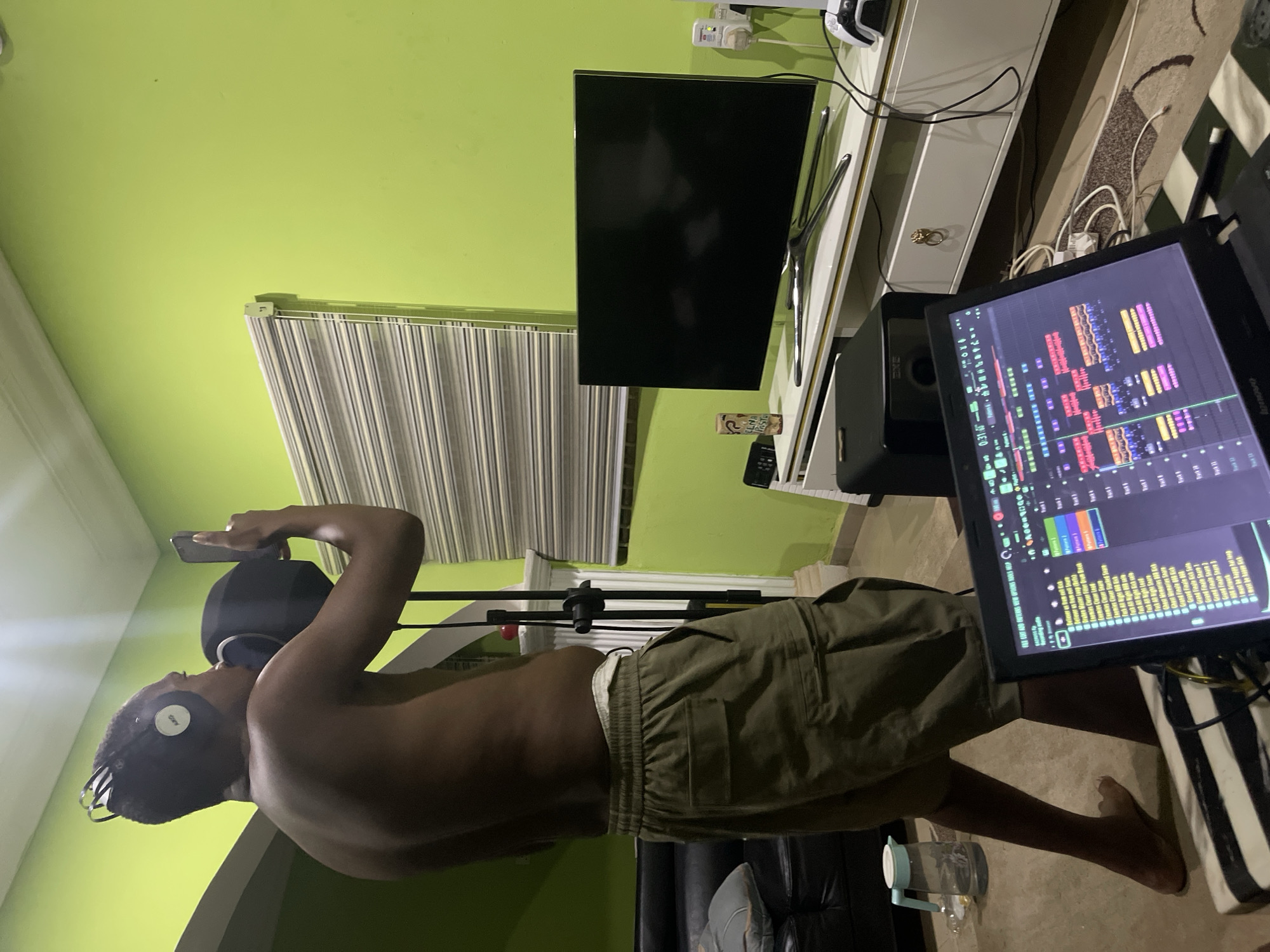 A man stands shirtless, wearing khaki shorts and headphones, in what appears to be a home recording studio. He is holding a phone near a microphone. A computer screen displaying audio editing software is visible to the right. The room has green walls, a window with blinds, a TV and a white cabinet against the wall. There is a water bottle on the floor near a black leather couch. The image does not provide information about the location being in Abuja, Nigeria.