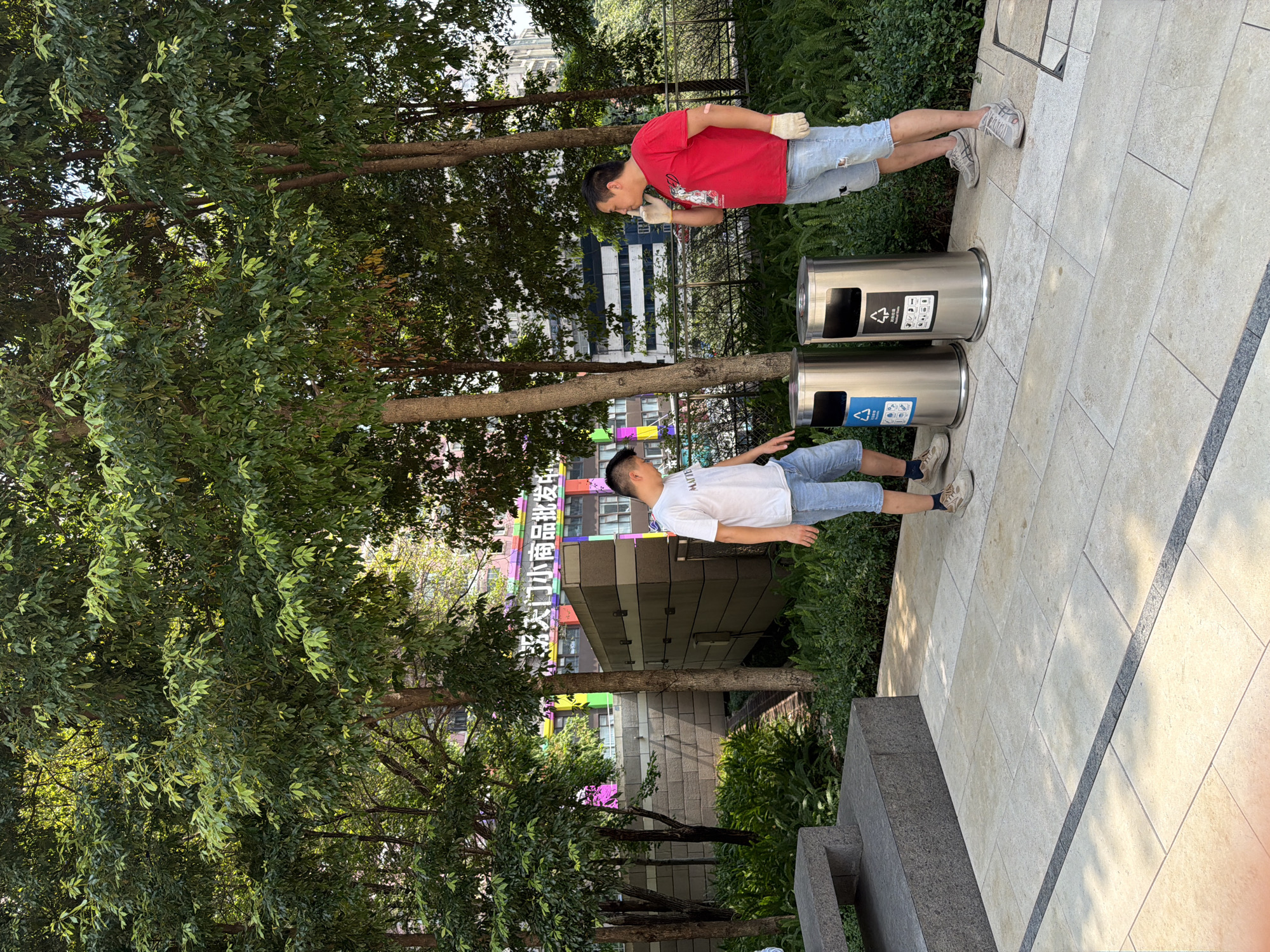 Two men are standing near two silver trash cans in an outdoor urban environment, likely in Yuzhong District, China. The man on the left, wearing a white t-shirt and denim shorts, is looking down at the trash cans. The man on the right, wearing a red t-shirt and distressed denim shorts, is holding his hand to his mouth as if speaking on a phone, and wearing white gloves. Behind them are lush green trees, dense foliage, and a building with colorful signage. The ground is paved with large light-colored tiles.