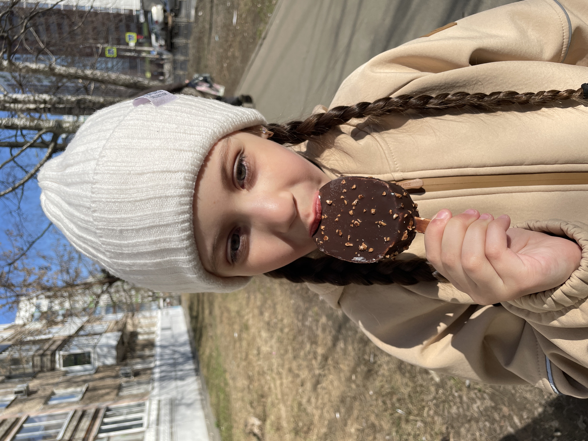 A young girl with fair skin, light eyes, and braided dark hair is holding and eating a chocolate-covered ice cream bar with nuts. She is wearing a white knit hat and a tan-colored jacket. The background is a blurred cityscape with trees and buildings. The location is likely Izhevsk, Russia.