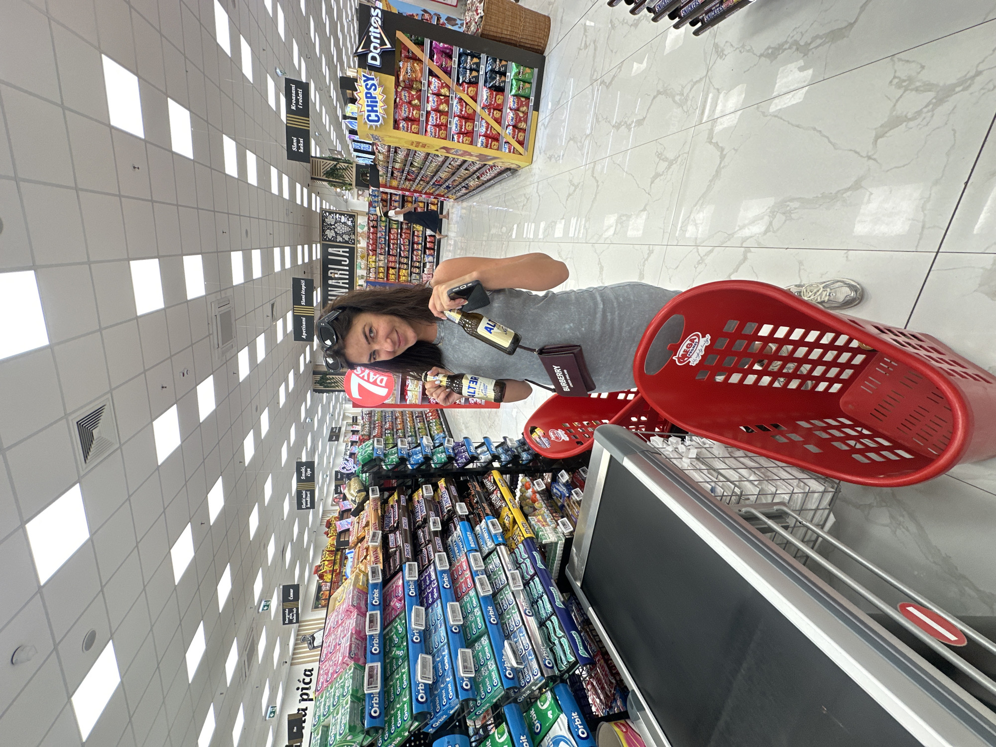 The image captures a woman in a supermarket in Budva, Montenegro. She is standing near a checkout counter and a red shopping cart, holding two bottles of beverages. The woman is wearing a gray dress and sunglasses on her head. The supermarket aisle displays shelves stocked with various confectionery and snack items, including Orbit gum, Snickers, Bounty, and Doritos. The environment appears to be well-lit with a modern ceiling. The floor is tiled with a marble pattern. In the background, other shoppers can be seen browsing the aisles, and there are various signs and displays typical of a retail setting.