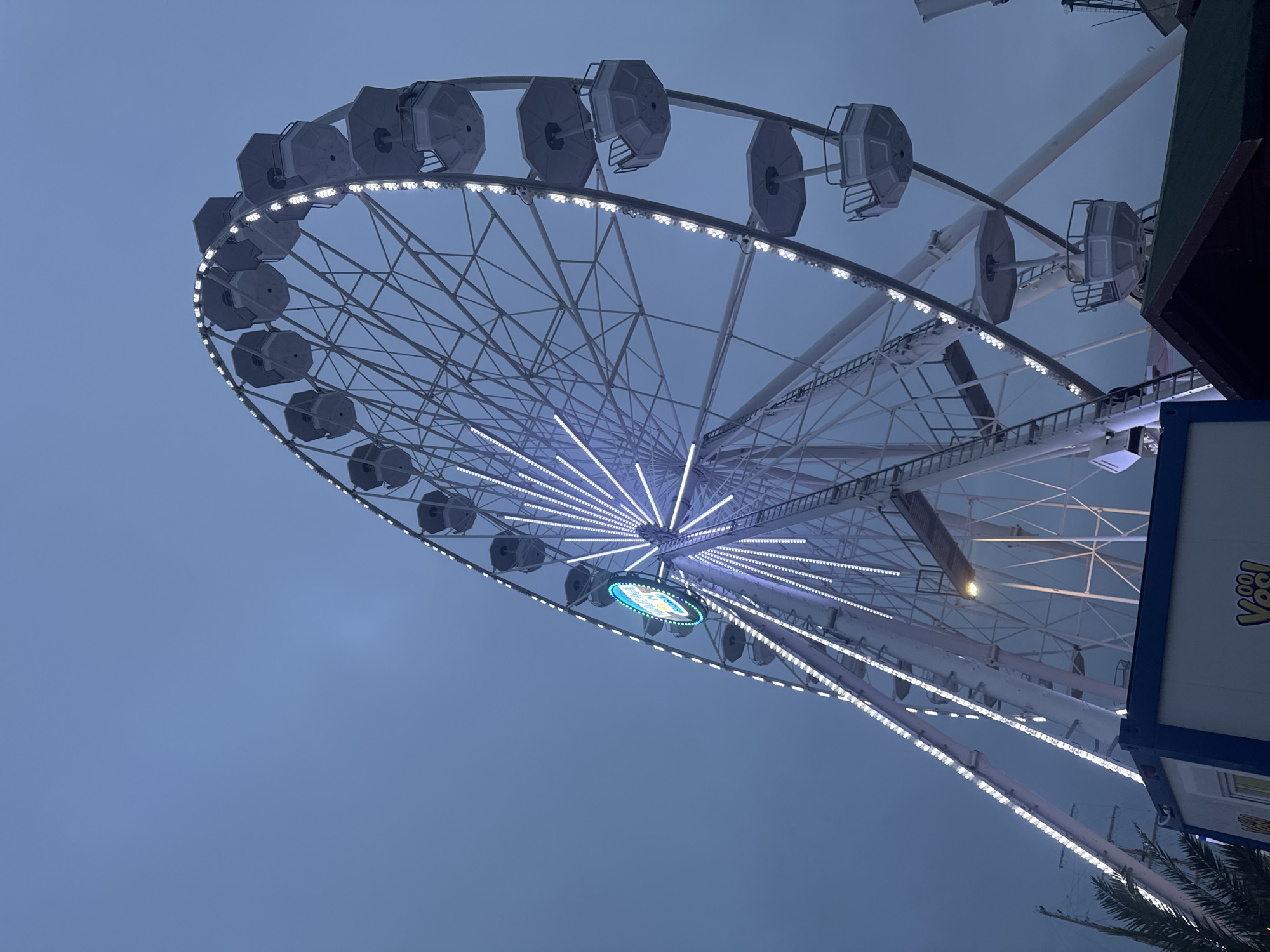 A large Ferris wheel, illuminated with white LED lights, stands against a dark, overcast blue-grey sky at dusk or night. The wheel's white metal frame is outlined by numerous bright white lights along its circumference and spokes, creating radial patterns. A circular, internally lit sign at the hub of the wheel displays blue and white illumination, featuring a logo that appears to depict a cityscape or landmark, likely indicating its location in Varna, Bulgaria. Approximately 20-25 light-grey, octagonal gondolas are visible, some appearing to contain occupants, positioned around the wheel's perimeter.
At the bottom left, the fronds of a palm tree are partially visible. In the central foreground, a white and blue rectangular structure, resembling a kiosk or container, features yellow text that partially reads "Voo! Yes!". To the right, part of a building with a dark-colored roof is visible. The base structure of the Ferris wheel is discernable, with one yellow lamp emitting light. The scene is located in the city of Varna, Bulgaria, consistent with the Varna Eye Ferris wheel commonly situated near the port or Marine Garden. No specific actions or interactions are clearly depicted.