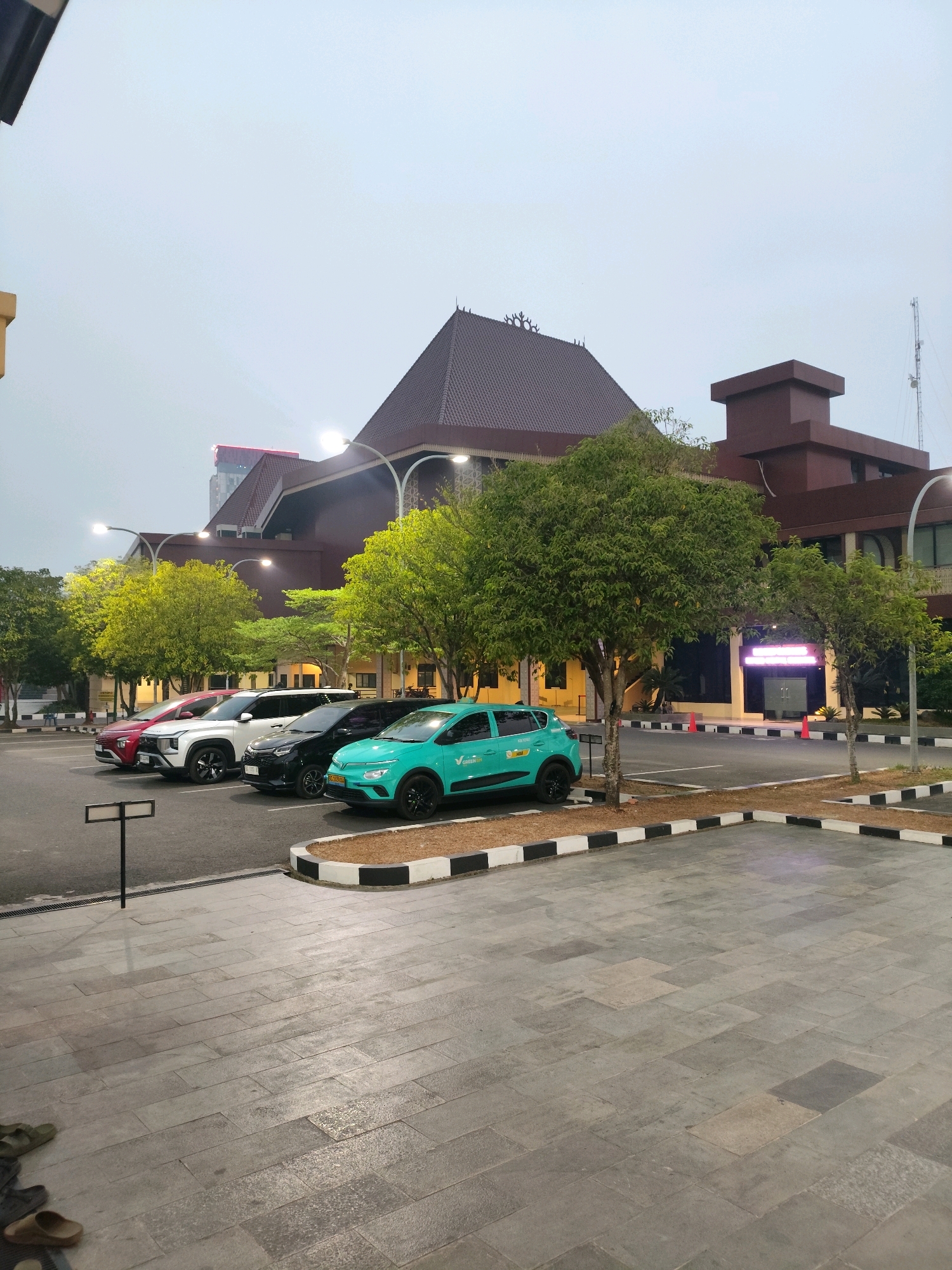 The image depicts a parking lot in front of a large, modern building with a traditional Indonesian roof. Four cars are parked in the lot, including a red SUV, a white SUV, a black SUV, and a teal electric car. The teal car has "GOJEK" written on its side, indicating it is likely a ride-sharing vehicle. The building appears to be a government or public institution, possibly a museum or a cultural center, given its architecture. A prominent neon sign is visible on the front of the building, but its text is illegible. The scene is illuminated by streetlights, and the sky is overcast, suggesting it is either dawn or dusk. The ground in the foreground is paved with grey tiles, and a few pairs of sandals are scattered on the ground. The overall impression is one of a well-maintained urban environment. This location is likely the Griya Agung, the official residence of the Governor of South Sumatra, which is located in Palembang, Indonesia.