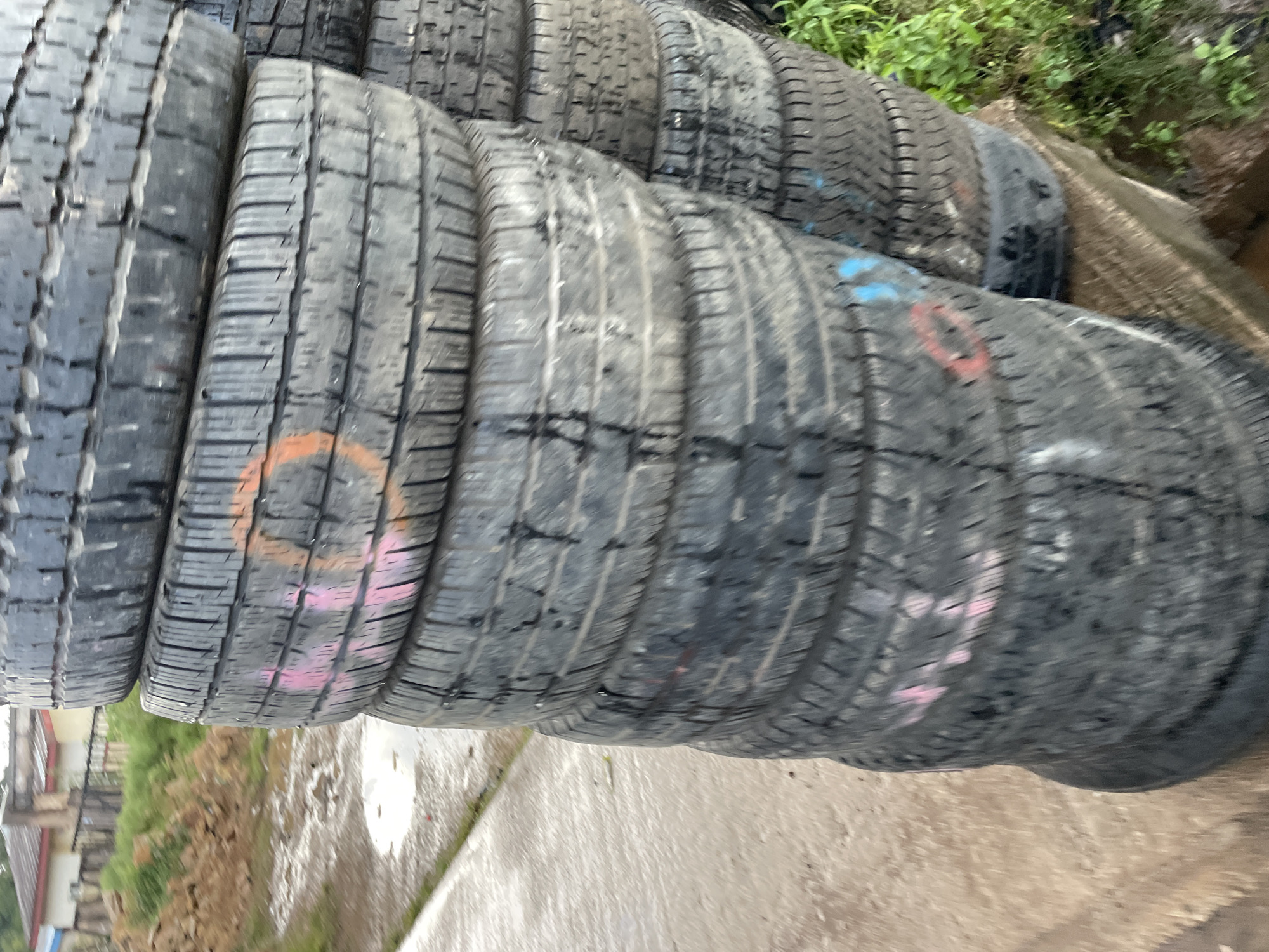 A stack of used tires is visible in a yard in Barakin Lerawi, Nigeria. The tires are black and appear to be made of rubber. They are stacked one on top of another, forming a tall tower. Some of the tires have markings on them, including orange, pink, and blue paint. The ground around the tires is wet and appears to be dirt or mud. In the background, there is a building with a red roof, some green vegetation, and a pile of brown dirt. The overall scene suggests an outdoor storage area for old tires.