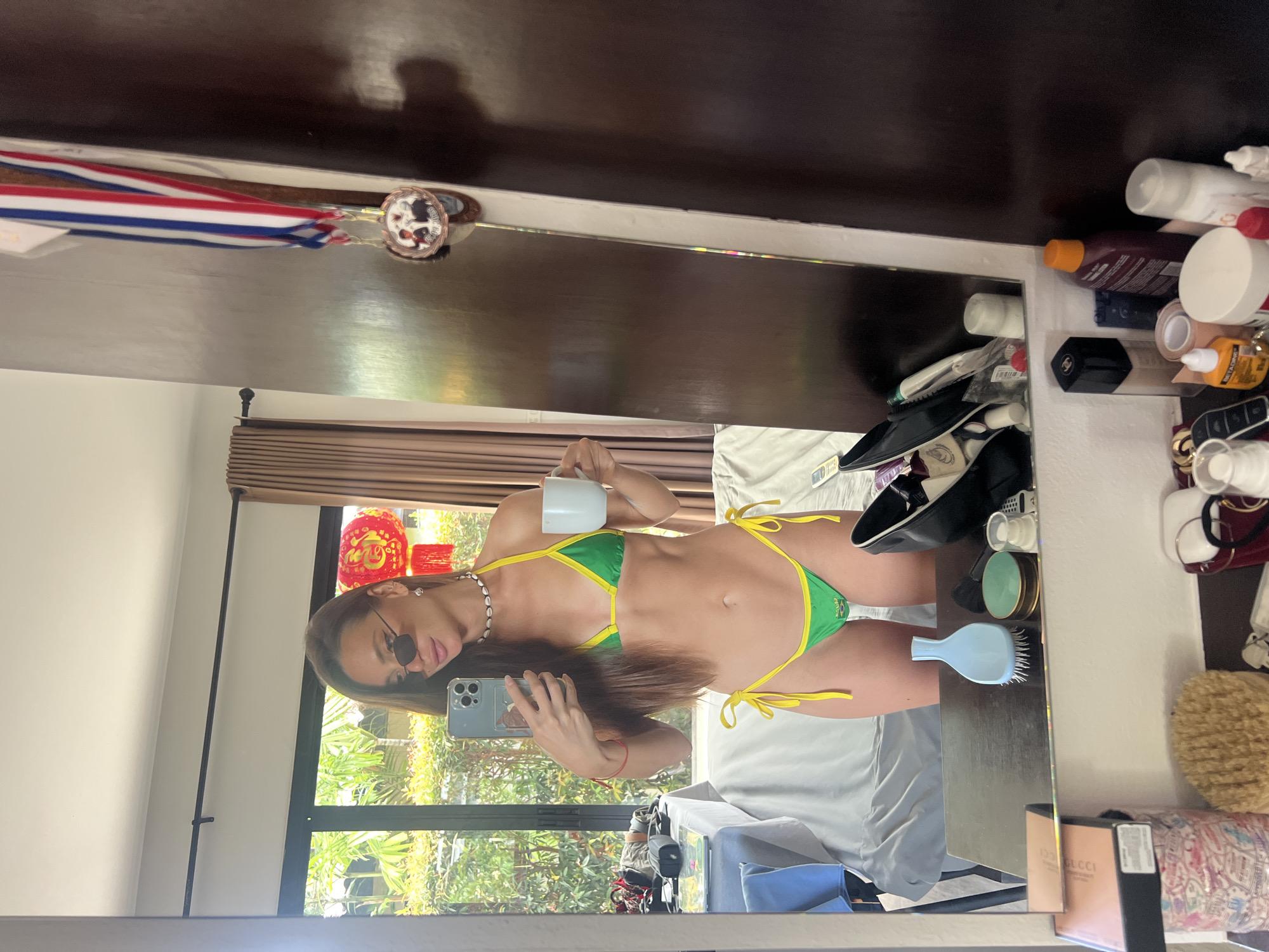 A woman stands in front of a mirror, taking a selfie. She is wearing a green and yellow bikini and sunglasses, and holding a white mug. In the background, there is a window with lush greenery visible outside, and two red lanterns hanging. The room appears to be a bedroom, with a bed in the background and a dresser in the foreground. The dresser is cluttered with various items, including makeup, toiletries, and a hairbrush. On the right side of the image, there is a closet door with medals hanging on it. The overall lighting suggests daytime.