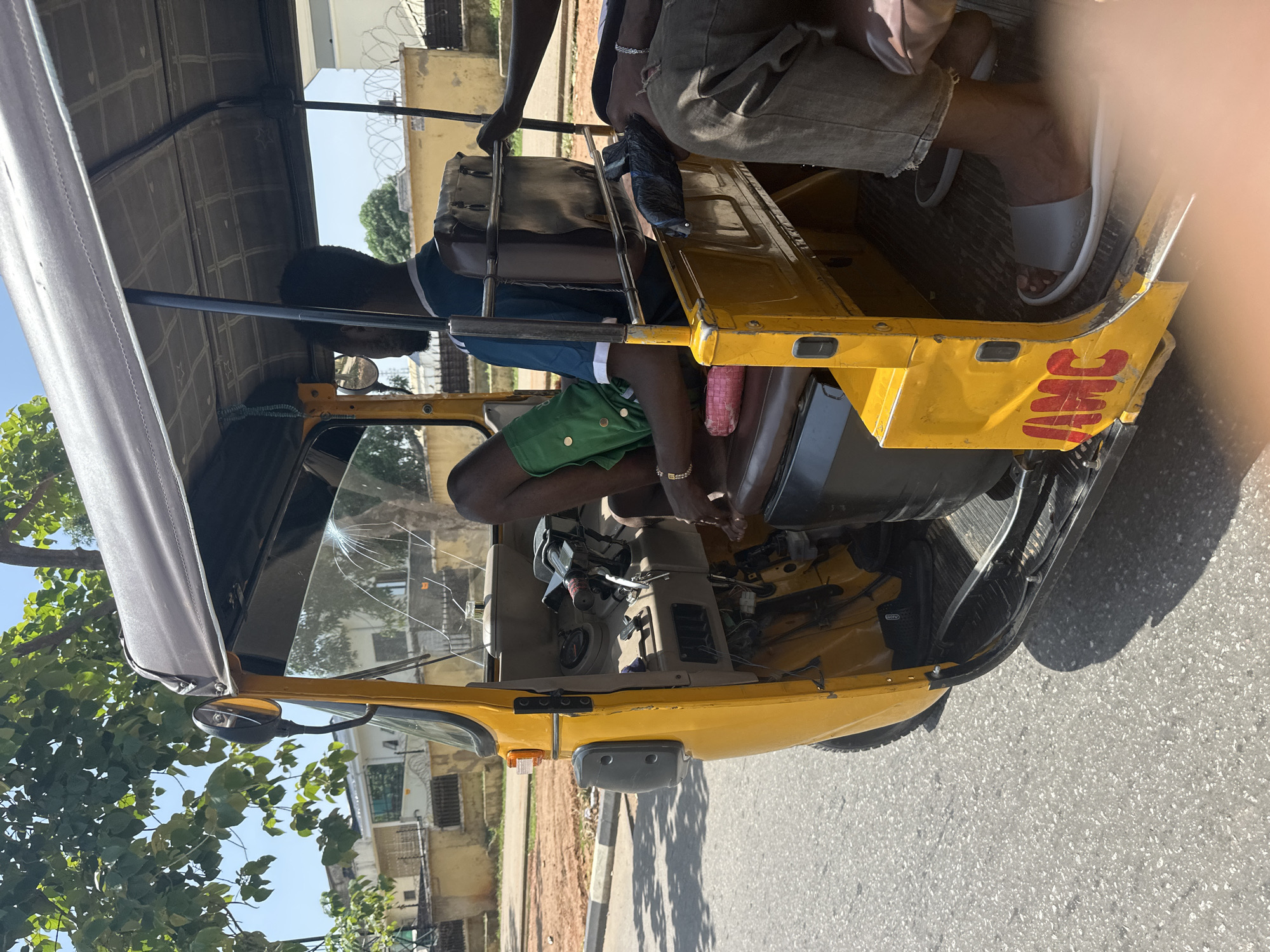 In the city of Abuja, Nigeria, a yellow auto-rickshaw, also known as a Keke Napep, is captured in motion on a paved road under a clear, bright sky. The driver, a Black male wearing a blue and white t-shirt and green shorts, is seated at the controls. He has a gold bracelet on his left wrist. A passenger, whose legs and feet are visible, is seated to the right of the driver, wearing ripped grey jeans and white slides. The auto-rickshaw has a dark canvas roof supported by metal poles, and the windshield is cracked. The interior features brown leather seats and a beige dashboard. The exterior of the auto-rickshaw has the word "AMC" painted in red on its side. The surrounding environment includes buildings and trees, with a barbed-wire fence visible in the background.