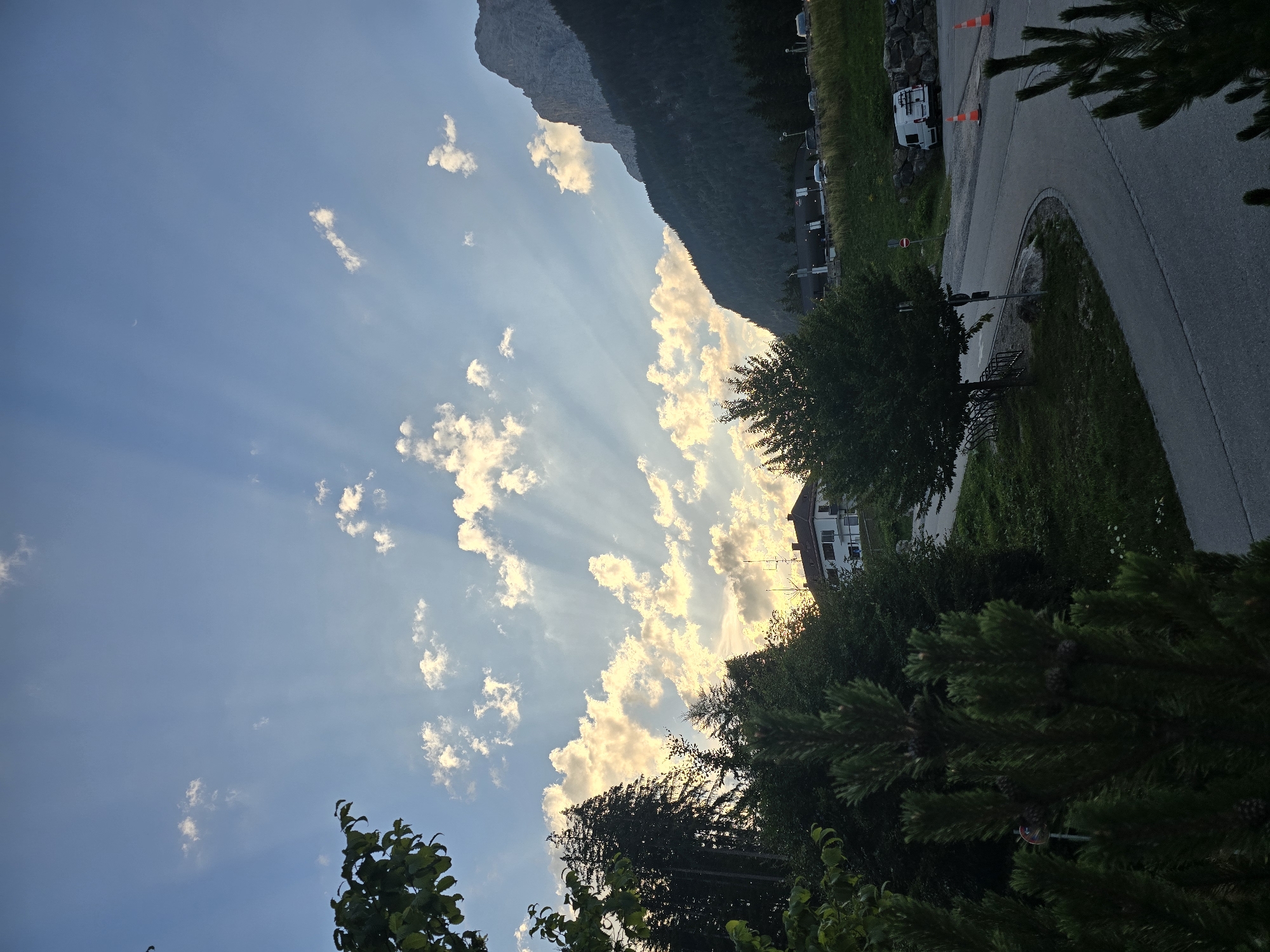 In the town of Grainau, Germany, a serene outdoor scene unfolds under a sky painted with crepuscular rays. Sunlight streams through scattered clouds, casting ethereal beams across the blue expanse. In the middle ground, a white building with a red-tiled roof sits atop a grassy incline, surrounded by lush green trees. To the right, the imposing silhouette of a dark, forested mountain range dominates the horizon, its peak partially obscured by shadows. A paved road winds through the foreground, bordered by a grassy verge. A white camper van is parked on the side of the road, accompanied by two bright orange traffic cones. Several dark green coniferous trees stand tall, their branches reaching towards the sky, adding a sense of depth and natural beauty to the landscape.