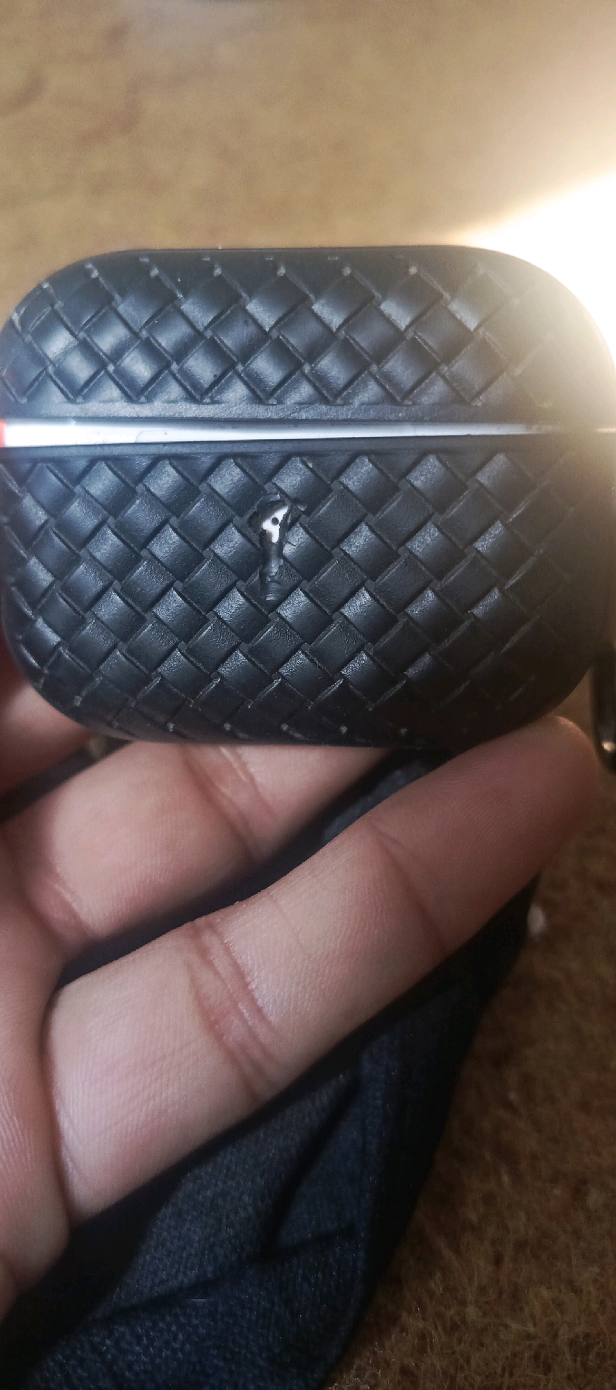 The image shows a close-up of a black, woven textured case for what appears to be wireless earbuds. The case has a metallic silver strip across the top. There is a small, dark hole with white material visible inside on the front of the case. A human hand, with visible fingerprints and nail beds, is holding the case, and a dark fabric material is visible beneath the case and hand. The background is a brown, textured surface, likely a carpet or wood. The scene appears to be indoors. There is no discernible information within the image to associate it with the city of Miana Thub, Pakistan.