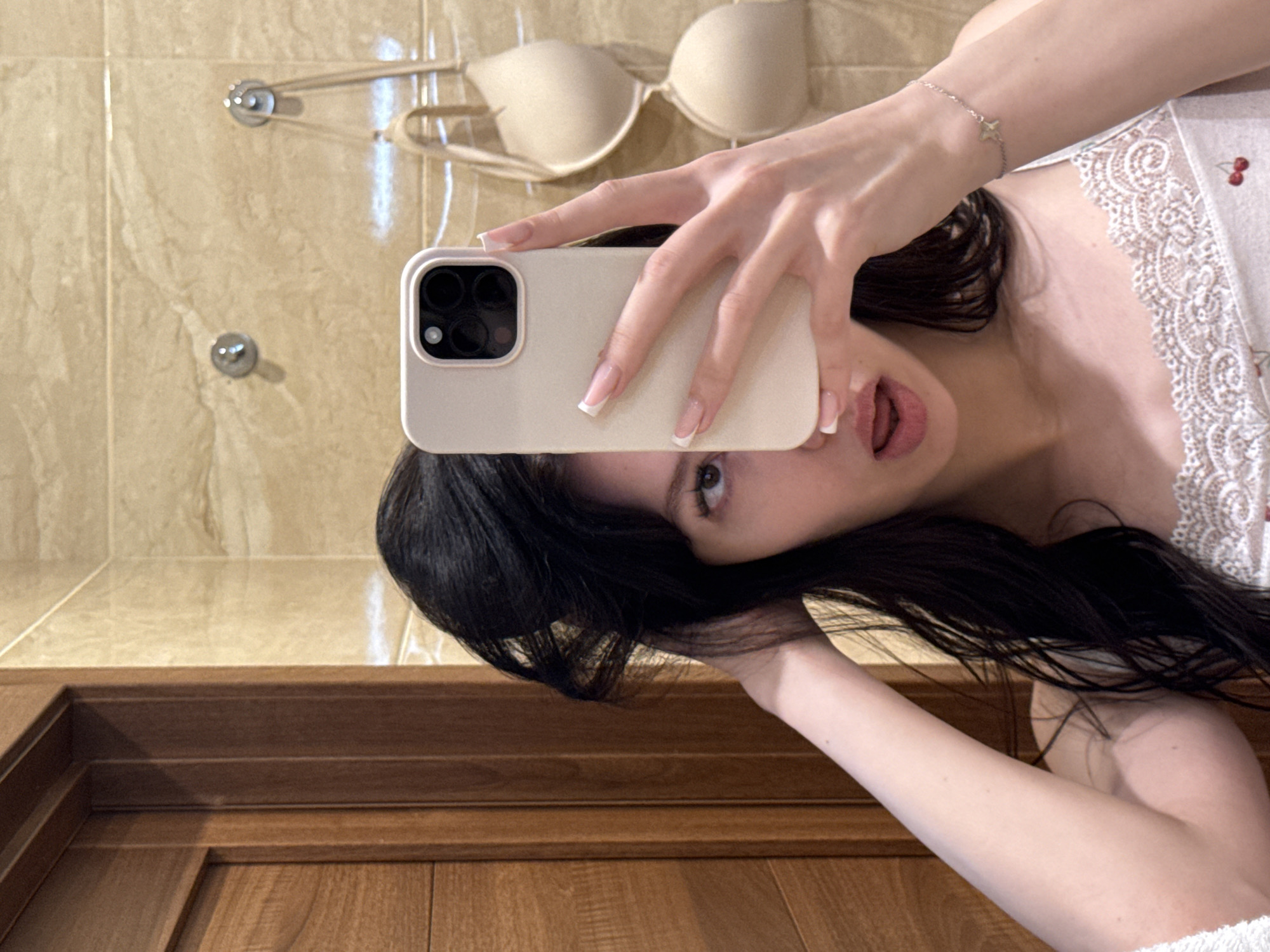 The image shows a young woman taking a mirror selfie in what appears to be a bathroom. She has long, dark hair that she is pulling up with her left hand. She is wearing a white camisole with lace trim and a cherry pattern. Her right hand is holding a light-colored smartphone, also partially obscuring her face. Her lips are slightly parted. In the background, there is a tiled wall, a wooden doorframe to the left, and a beige bra hanging from a hook on the wall. The overall scene suggests a private, personal moment captured in a domestic setting. There is no information within the image to suggest a specific location in the city of Khimki, Russia.