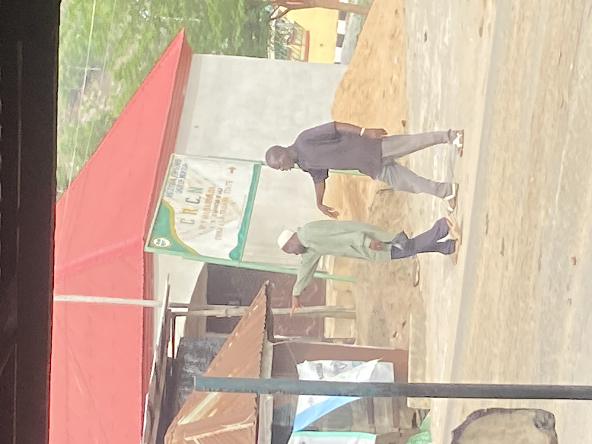 The image depicts two men standing outdoors in what appears to be Mararaba, Nigeria. One man is wearing a dark purple shirt and light-colored pants, and the other is wearing a light green long-sleeved shirt and dark pants with a white cap. Both are wearing sandals. They appear to be examining something on the ground, perhaps pointing or indicating something with their hands.
In the background, there is a building with a red roof and a sign for CRCN (presumably an organization). There is also a pile of sand and some other structures with corrugated metal roofs. The scene is generally cluttered, suggesting a busy or developing area. The ground is a light-colored, concrete surface.