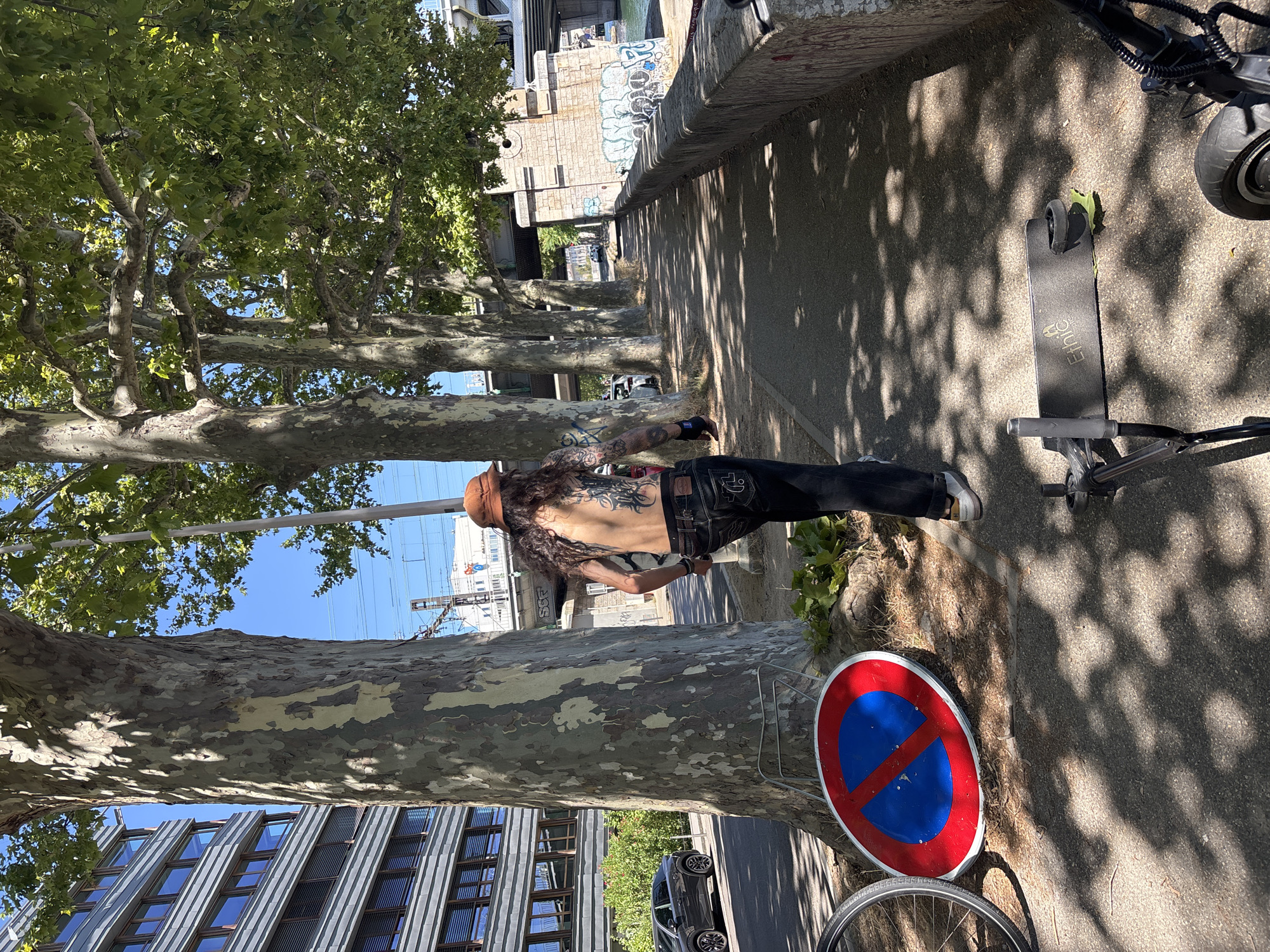 The image captures a section of the Quai Saint-Vincent in Lyon, France, on a sunny day. A shirtless person with extensive tattoos on their back and arms, wearing a brown hat, dark jeans, and sneakers, is walking away from the camera on a paved walkway. In the foreground, a red and blue 'no parking' sign is lying on the ground next to a bicycle wheel. Further along the walkway, two electric scooters are visible. The walkway is bordered by a stone wall on the right, and large, mature trees with dappled shade line the left side of the path. In the background, a modern building with horizontal bands of windows and a bridge over the Saône River can be seen.