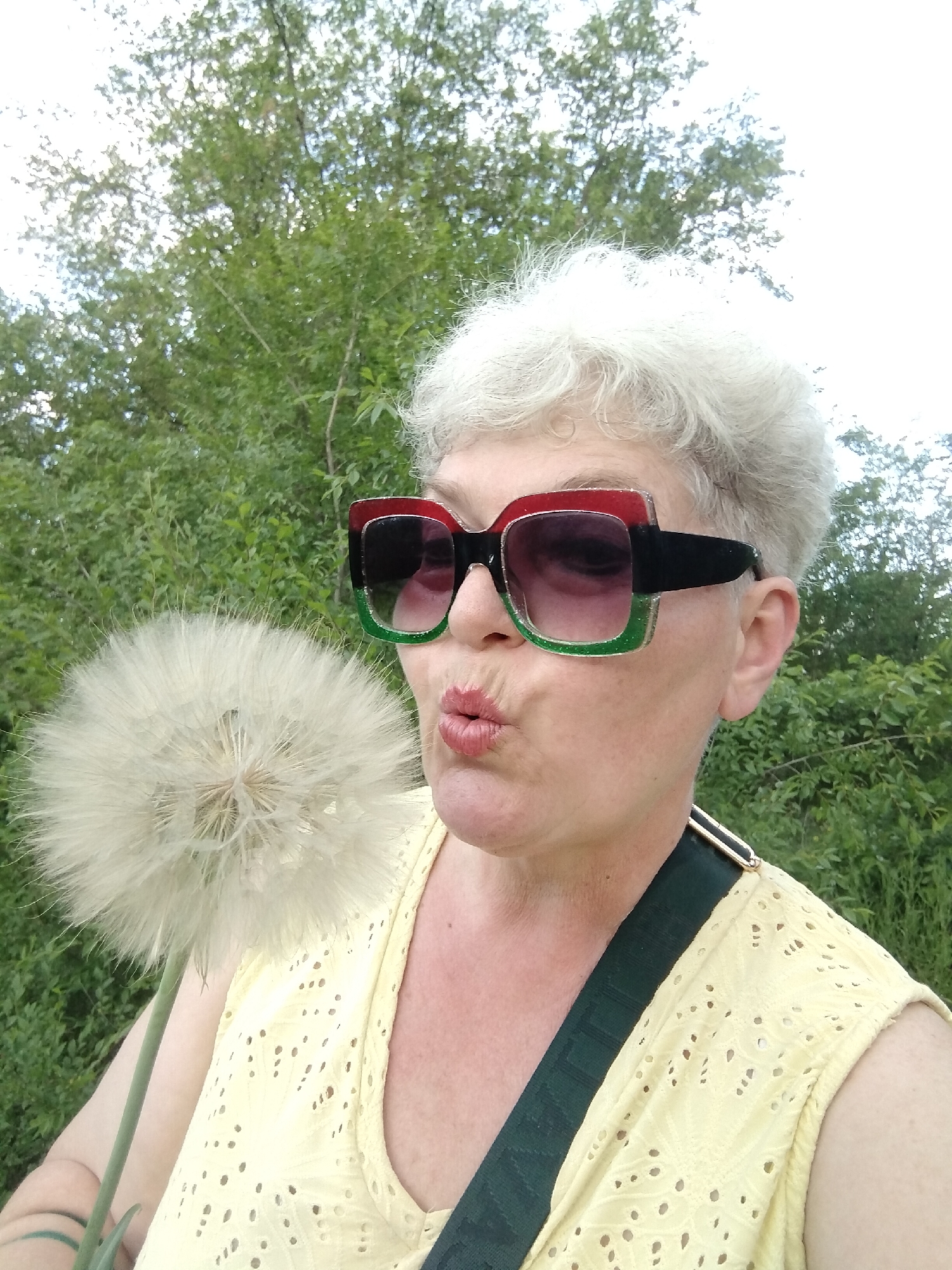 An outdoor, eye-level photograph, taken in a selfie-style, captures a person of apparent female gender in Horishni Plavni, Ukraine. The subject is depicted from the upper torso upwards, with the right side of their face and head prominent. They have short, curly, light grey hair. The subject is wearing large, square-framed sunglasses with a thick black frame; the upper left portion of the frame is red and glittered, while the lower left portion is green and glittered. The lenses are dark and appear to have a subtle gradient. The subject's lips are pursed in a kissing or blowing gesture. They are wearing a light yellow sleeveless top featuring eyelet cutouts. A dark green fabric shoulder strap, possibly from a bag, with a gold or metallic buckle-like element, is visible over the subject's right shoulder. In the foreground, on the left, the subject holds a very large, spherical dandelion seed head (Tragopogon-like, also known as giant salsify) with a visible green stem. The seed head is white and fluffy, composed of numerous pappus structures. The background consists of dense green foliage, consistent with trees and shrubs, under a bright, overcast, or hazy white sky.