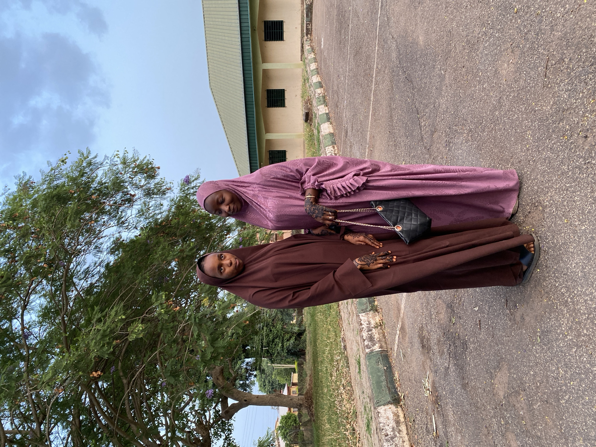 Two women stand outdoors in Vom, Nigeria, on an asphalt surface, likely a road or parking area. A light green building is in the background. One woman is wearing a brown hijab and gown, and the other is wearing a light purple hijab and gown and carrying a black quilted handbag with a gold chain. Both women have henna designs on their hands. A tree with green leaves and purple flowers is visible to the left. The sky is light blue. The scene appears to be during the day.