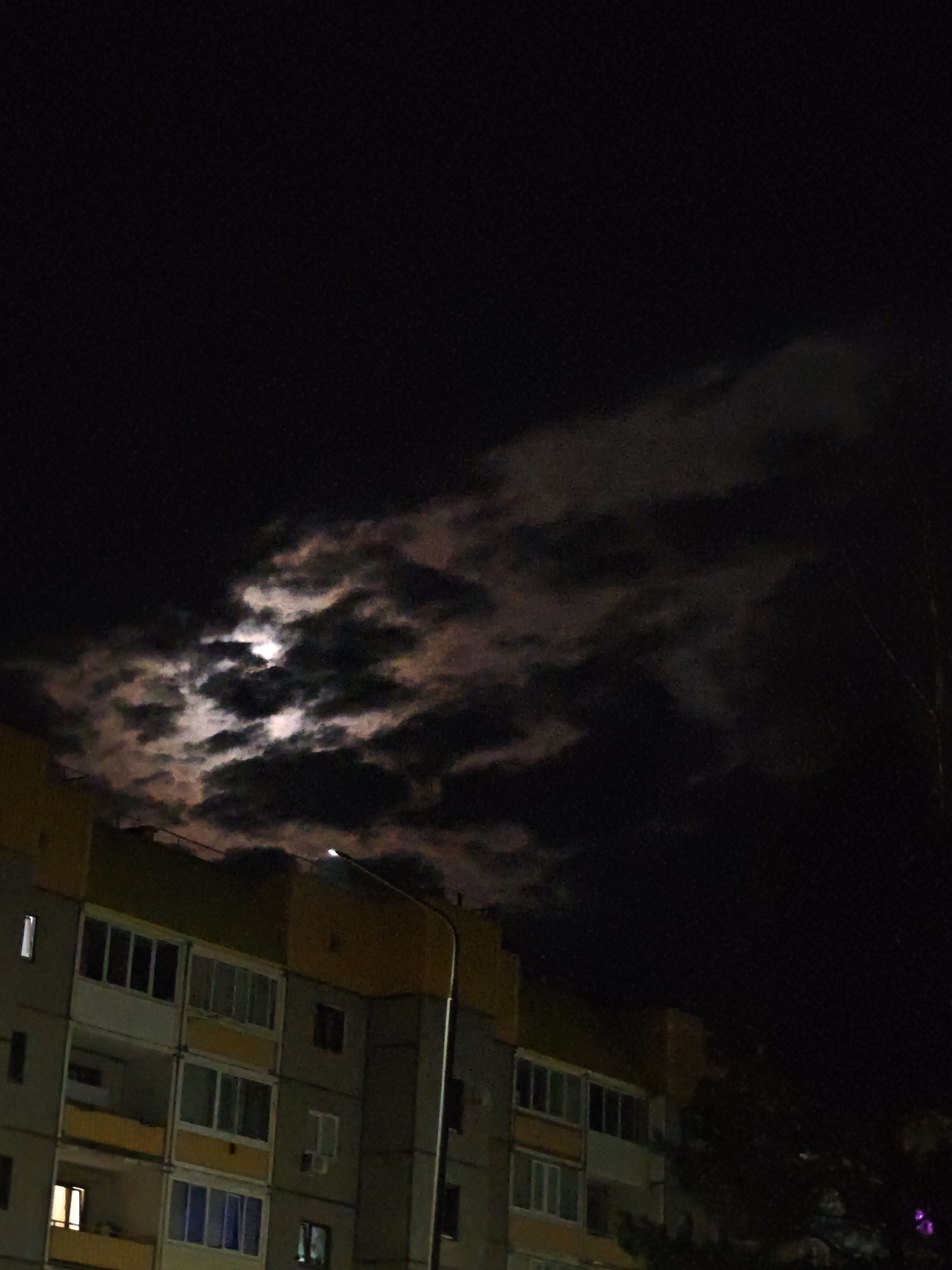 A nighttime image shows an apartment building, possibly located in the city of Марковский, Russia, as indicated in the prompt. The building is several stories tall with lit windows and yellow accents. In the dark sky, clouds cover the moon, creating a dramatic effect. A street lamp stands in front of the building. A tree is partially visible in the lower right corner.
