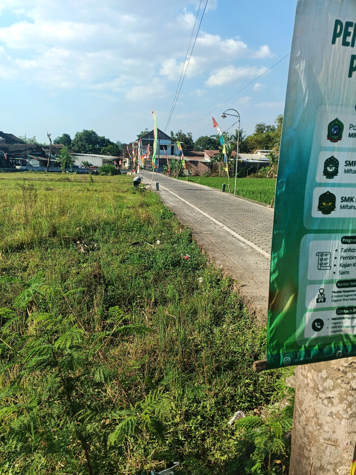 This is a medium shot, eye-level, outdoor photograph taken in the city of Ungaran, Indonesia. The scene depicts a paved road with decorative flags lining one side, leading into a village. To the left of the road is a grassy field. A large green banner with text is partially visible on the right side of the frame, attached to a wooden pole. The banner appears to be an advertisement for educational institutions, listing programs such as Tahfidz, Pembinaan Qiroah, Kajian Kitab Kuning, and Sains. It also provides contact information for Pondok Pesantren and its administration in Ungaran. The sky is clear with some clouds, and the overall environment suggests a sunny day.