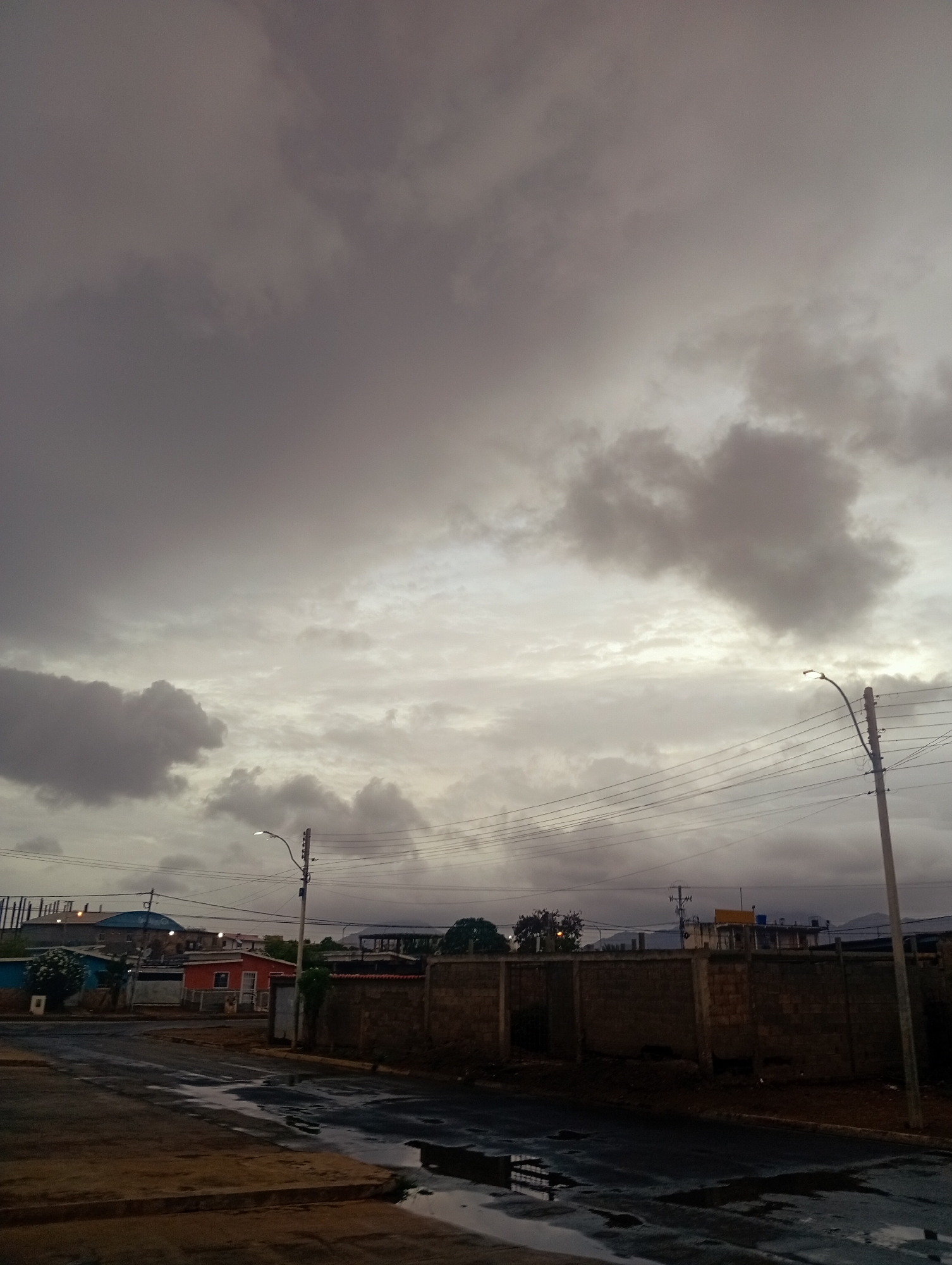 The image captures a street scene in Juan Griego, Venezuela, under a heavily overcast sky, suggesting impending rain or recent rainfall as indicated by wet patches on the asphalt. The foreground shows a street corner with dark asphalt and puddles reflecting the sky. To the right, a tall utility pole with numerous wires stretches upwards. Along the street, a brick wall runs horizontally, separating the road from the properties behind it. Various buildings are visible in the background, including a small orange house with a white trim, and other structures with colorful roofs and facades, some appearing to be under construction with scaffolding. The overall atmosphere is somber and muted due to the cloudy conditions. There are no people or vehicles visible in the frame.