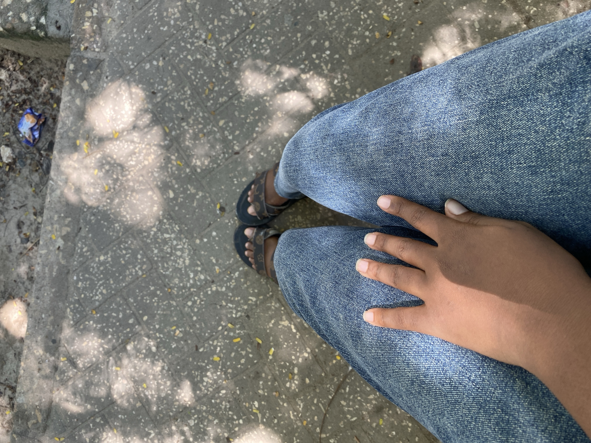 A photograph taken from an elevated perspective shows a person sitting on an outdoor ledge or step, likely in an urban environment. The individual is wearing blue denim jeans and dark-colored sandals. Their hand rests on their lap. The ground, possibly a sidewalk, is paved with gray interlocking stones, showing mottled lighting, likely from sunlight filtering through trees. A piece of discarded blue and silver trash lies near the edge of the frame, on the left. It is not possible to confirm the location in Dar es-Salaam, Tanzania, based on the visual information provided.