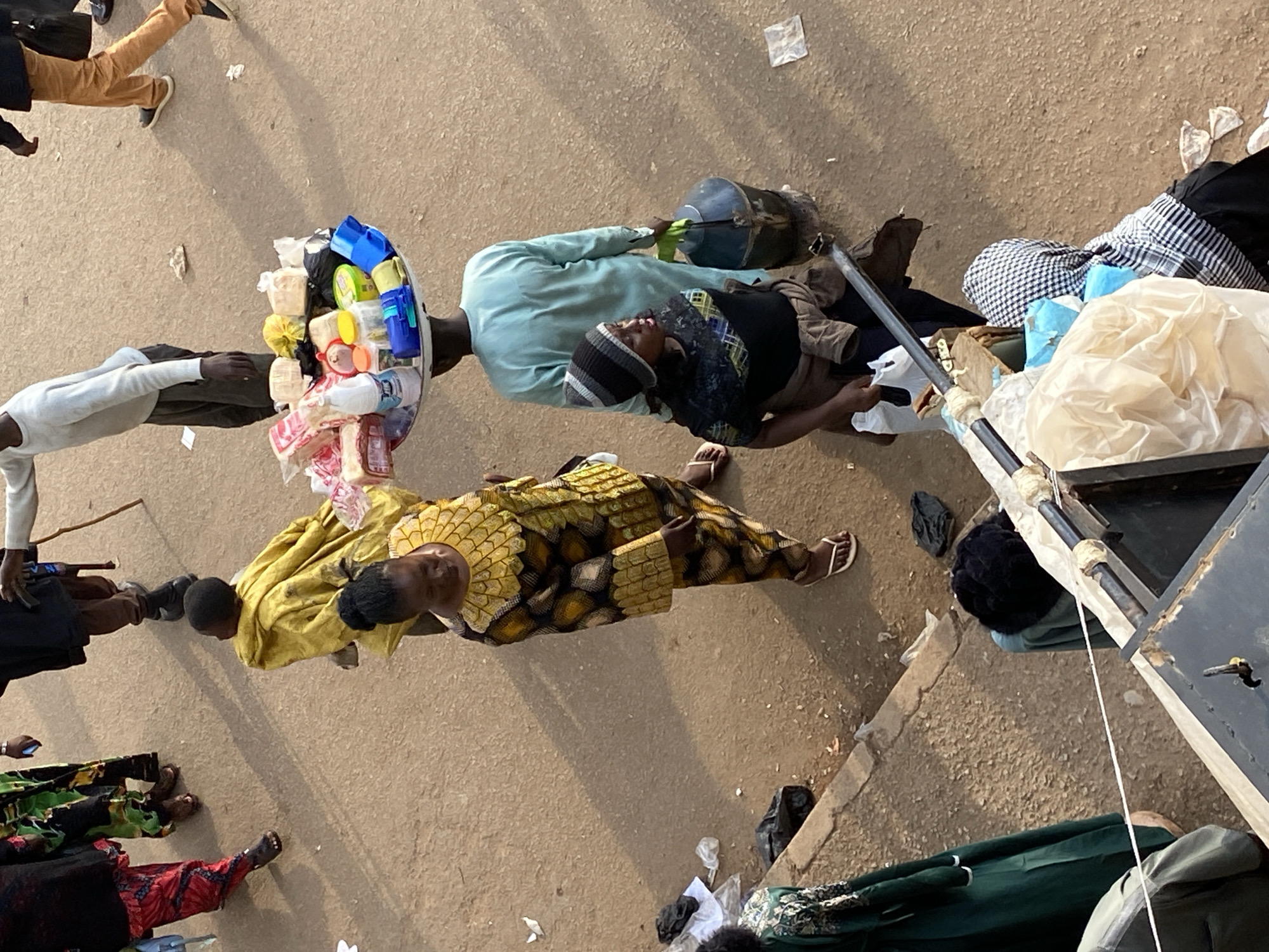 This image captures an overhead view of a busy street scene in Bukuru, Nigeria, under daylight conditions. The ground surface is a rough, light brown or tan asphalt or dirt, littered with various pieces of trash including plastic bags and paper scraps. Multiple individuals are present, engaged in various activities, with shadows cast to the left, indicating sunlight from the right. In the upper left quadrant, three individuals are partially visible, walking away from the viewer. One wears red patterned trousers and a dark top, with dark footwear. Adjacent to them is a person in a green patterned garment, also with dark footwear. A third person in dark clothing is partially obscured. In the upper right, two more individuals walk away. One wears a light gray long-sleeved shirt, dark pants, and dark shoes, holding a wooden stick in their right hand. Beside them, another person is seen in light brown/tan trousers and a dark jacket, wearing dark shoes. In the mid-ground, a young child, seen from behind, is draped in yellow fabric and appears to be looking to the left. Immediately behind the child, a woman wearing a gold and black patterned traditional outfit and white flip-flop sandals is looking downwards. Behind the woman, a man in a light blue long-sleeved garment, viewed from the back, balances a large, circular metal tray on his head. The tray contains a variety of items, including several packaged loaves of white bread, plastic bags containing yellow contents, small colorful plastic containers with lids, a white plastic bottle with a blue cap (possibly containing liquid), and other miscellaneous packaged goods. To the right of the man with the tray, a woman is seen facing slightly left. She wears a black long-sleeved top with a patterned design, dark pants, and a black, gray, and white striped beanie. She carries a large, dark, metallic pot with a green handle in her right hand, which appears to be resting on a small, dark base. In the lower left, a person in a dark green garment is partially visible, standing near a raised concrete curb. At the bottom center and right, a metal structure, possibly part of a market stall or cart, is partially visible. It has a dark frame and white, possibly plastic or fabric, covers. Items, including plastic bags, are resting on it. Behind this structure, a person is largely obscured, but a black and white