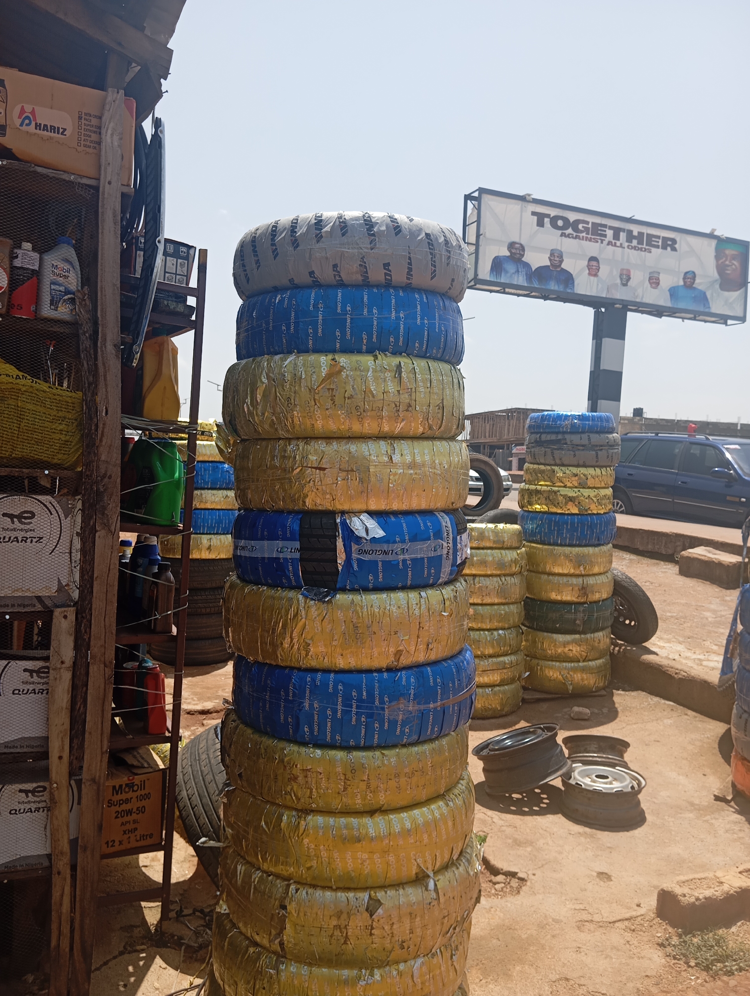 The image shows an outdoor tire shop. In the foreground, a tall stack of tires, wrapped in blue and gold packaging, dominates the scene. To the left, shelves are filled with various items, including boxes of "Hariz" and "Mobil Super" engine oil, as well as green and yellow canisters. Further back, a blue car is parked on a dirt lot. A billboard with the word "TOGETHER" and images of several men is visible in the background. The ground is a mixture of dirt and scattered debris, including several car rims. The overall impression is that of a busy, functioning commercial establishment in a potentially developing area.