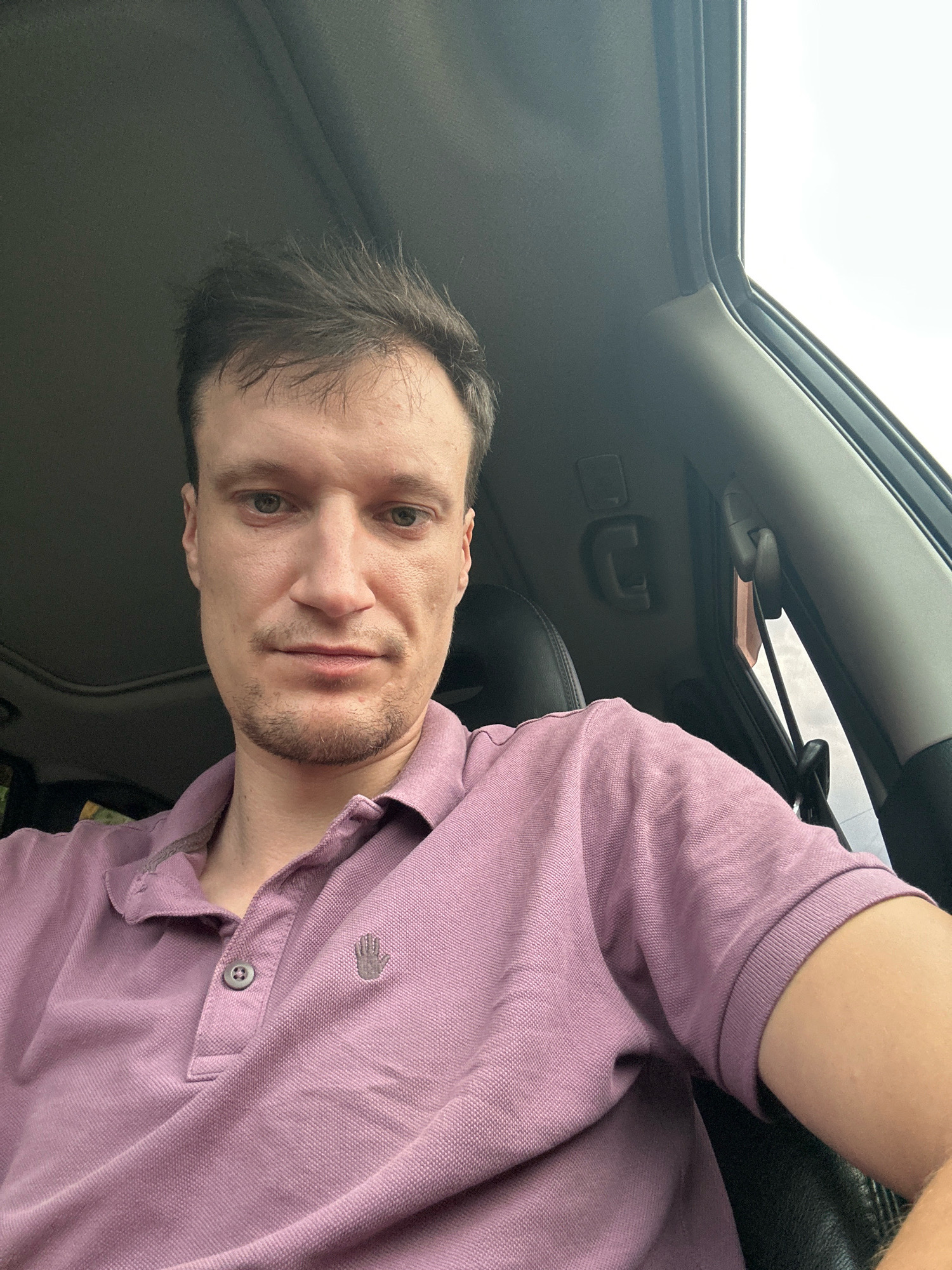 The image shows a man in the driver's seat of a car, looking at the camera. He is wearing a light purple polo shirt with a small hand logo on the left chest. The man has short, dark brown hair and a light beard. His expression is neutral.
The interior of the car is visible, including the dashboard and part of the passenger seat. There is a sunroof overhead, which is open, letting in natural light. The background outside the car window is bright, suggesting it is daytime. The weather appears to be overcast, as indicated by the diffused light.
The image was taken in Shatava, Ukraine. There is no visible text in the image. The setting appears to be a typical car interior, and the activity taking place is the man taking a selfie. The overall mood of the image is casual and everyday.