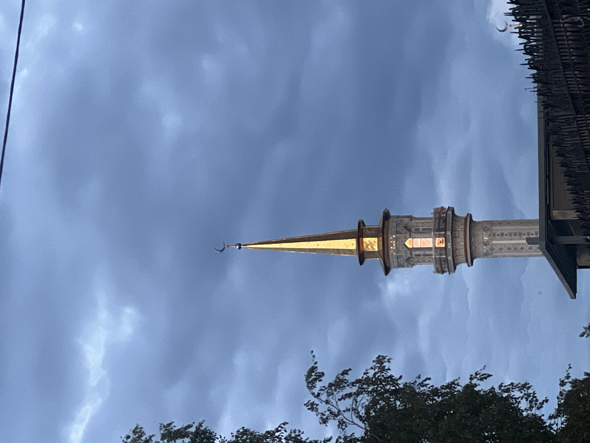 The image shows a minaret of a mosque in Surgut, Russia. The minaret is tall and slender, with a golden spire topped by a crescent moon. The lower part of the minaret is made of light-colored stone with decorative carvings. There is a window on the minaret that is lit from within. In the foreground, to the left, is a tree with dark green leaves. In the background, the sky is filled with dark, dramatic clouds, suggesting either dusk or an approaching storm. A dark metal fence with pointed spikes runs across the bottom of the image, obscuring the base of the mosque building. Another small crescent moon is visible above the fence in the distance.