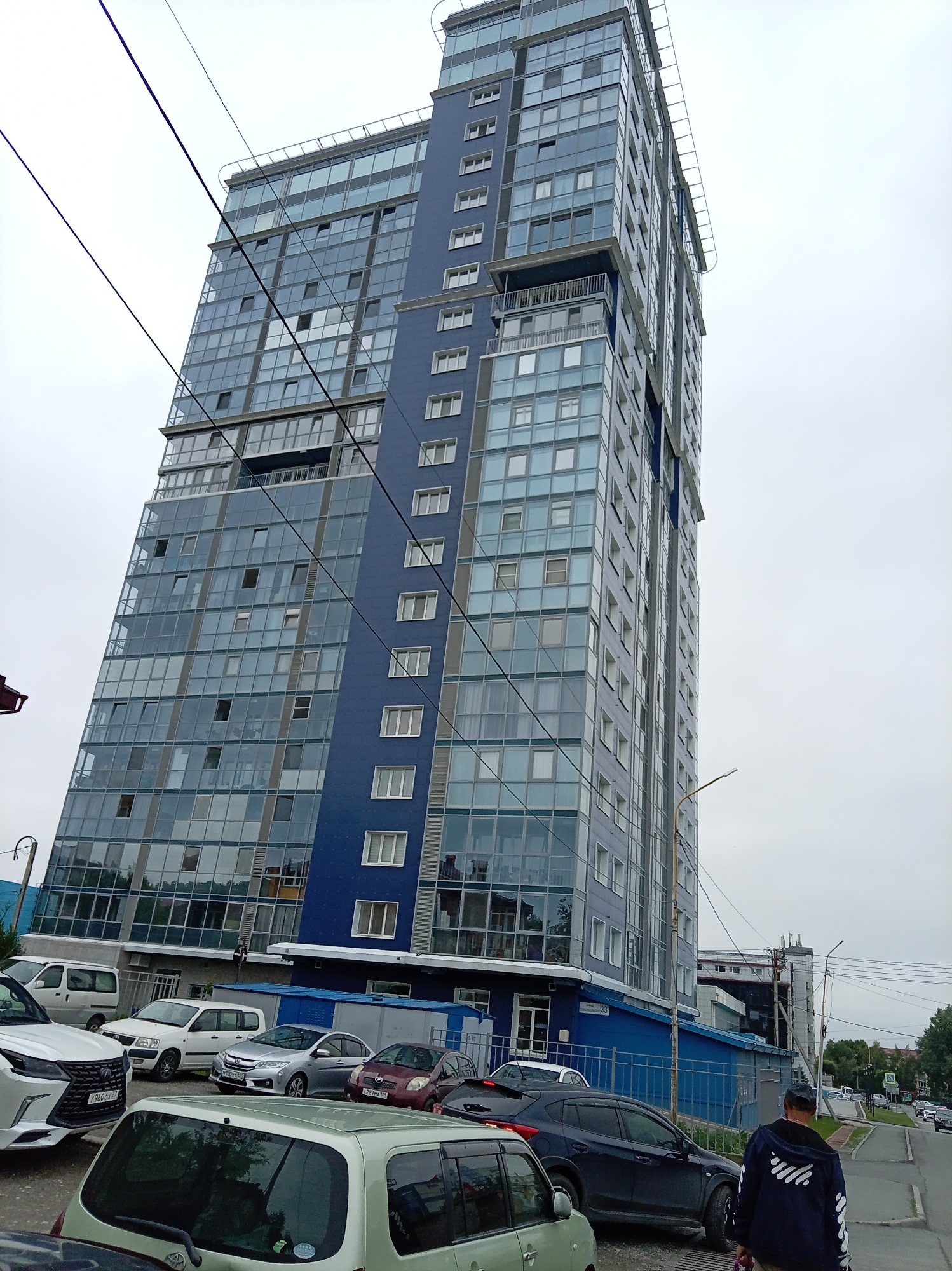In Artyom, Russia, a tall, modern blue and glass building stands under an overcast sky, angled towards the viewer. The building features numerous windows, some open, and a darker blue vertical section. In the foreground, several cars are parked: a white Lexus SUV with license plate Y960CX 27, a silver sedan with license plate H021TX 92, a maroon hatchback with license plate A287MA 125, and a light green minivan. A person wearing a dark hooded jacket with white stripes on the sleeves is walking away from the camera towards the right side of the frame. Power lines are visible crisscrossing the sky. A yellow street lamp is present near the building's base. The scene appears to be a residential or commercial area with paved surfaces and some greenery visible in the background.