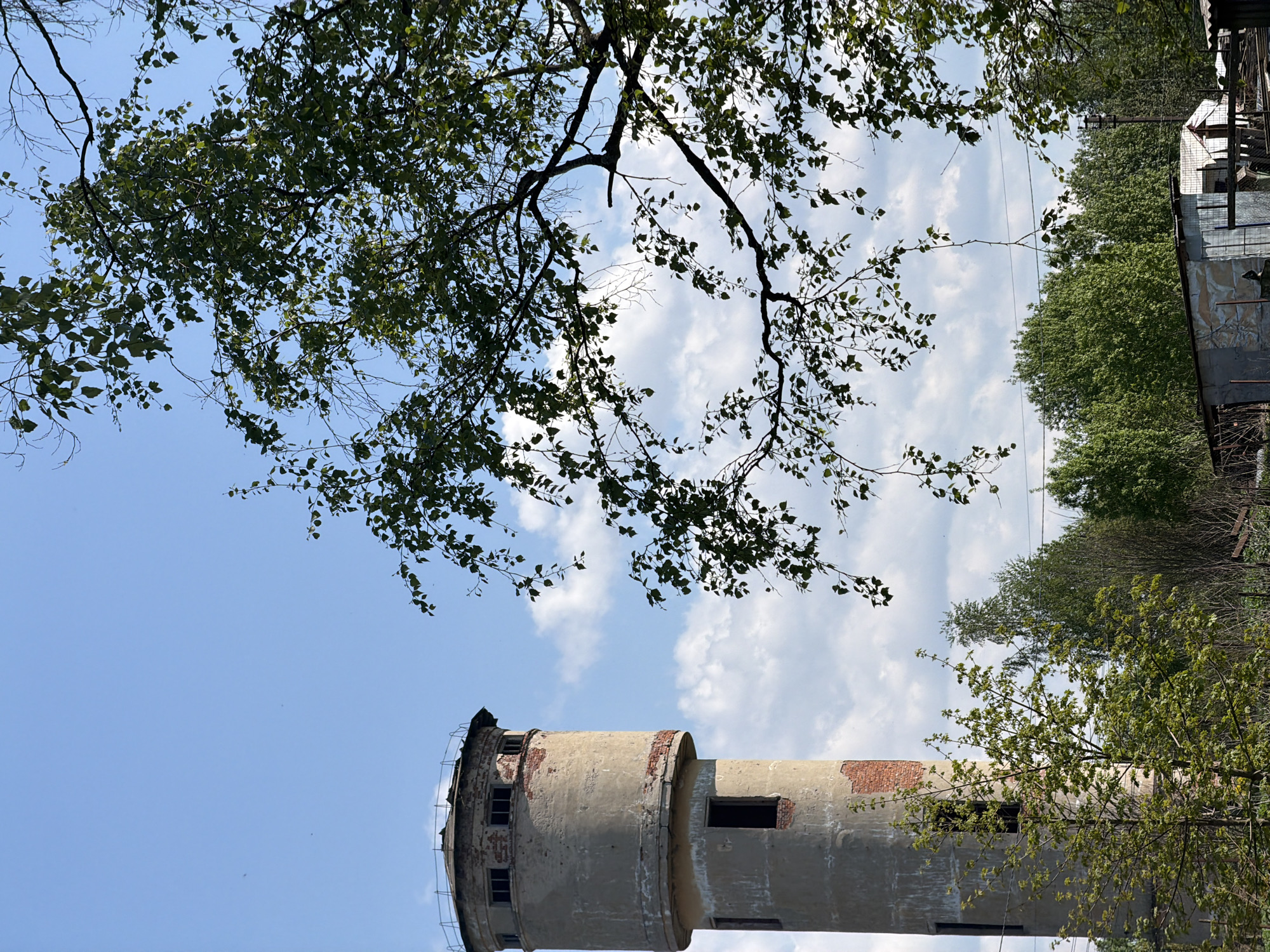 The image shows an old, cylindrical water tower in Tokkari, Russia. The tower is made of concrete and brick, with visible signs of decay and missing sections. It has small, empty window openings. Branches and leaves from nearby trees frame the tower against a blue sky with scattered white clouds. Other trees and a partially visible building are present in the background. The image is taken from a low angle, emphasizing the height of the tower.