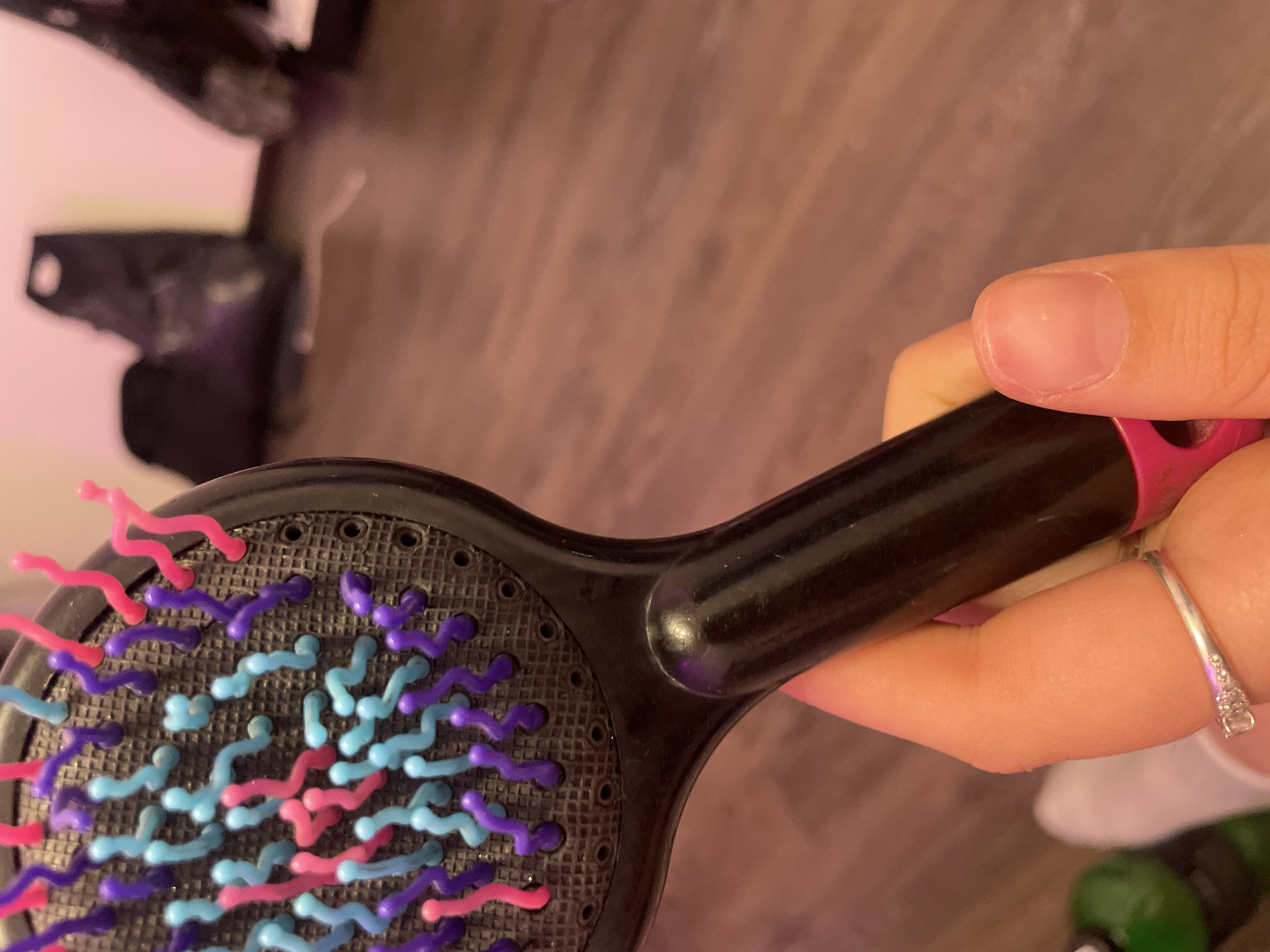 The image shows a close-up of a black hairbrush with colorful bristles in shades of pink, light blue, and purple. The brush is held by a hand with a silver ring on the index finger. The background is out of focus, showing a wooden floor and some objects that appear to be bags or clothing on the floor. The lighting suggests an indoor environment, possibly a room with pinkish ambient light. The image does not provide any information that would allow for the determination of its location in Kokkorevo, Russia.