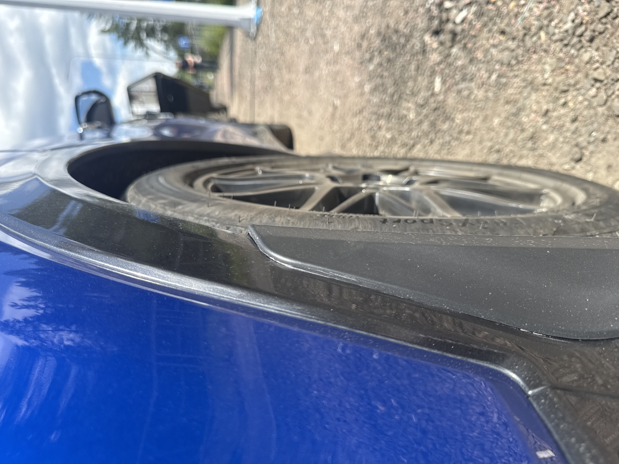 An extreme close-up shot focuses on the rear wheel and fender of a blue car, viewed from a low angle. The car's blue paint has reflections of trees. A black plastic fender flare is visible, with a mud flap attached below it. The wheel features a multi-spoke silver rim with a tire that has some visible lettering. The background is out of focus, showing a gravel path, a sidewalk with a white pole, and a second car with its door open. In the distance, a person in dark clothing walks away from the camera. The scene suggests an outdoor location, possibly a parking area or a road shoulder, with hints of a suburban or semi-rural environment given the presence of trees and a natural landscape. The specific location in Серебряный Бор, Russia, is implied by the context but not visually confirmed within the image itself.