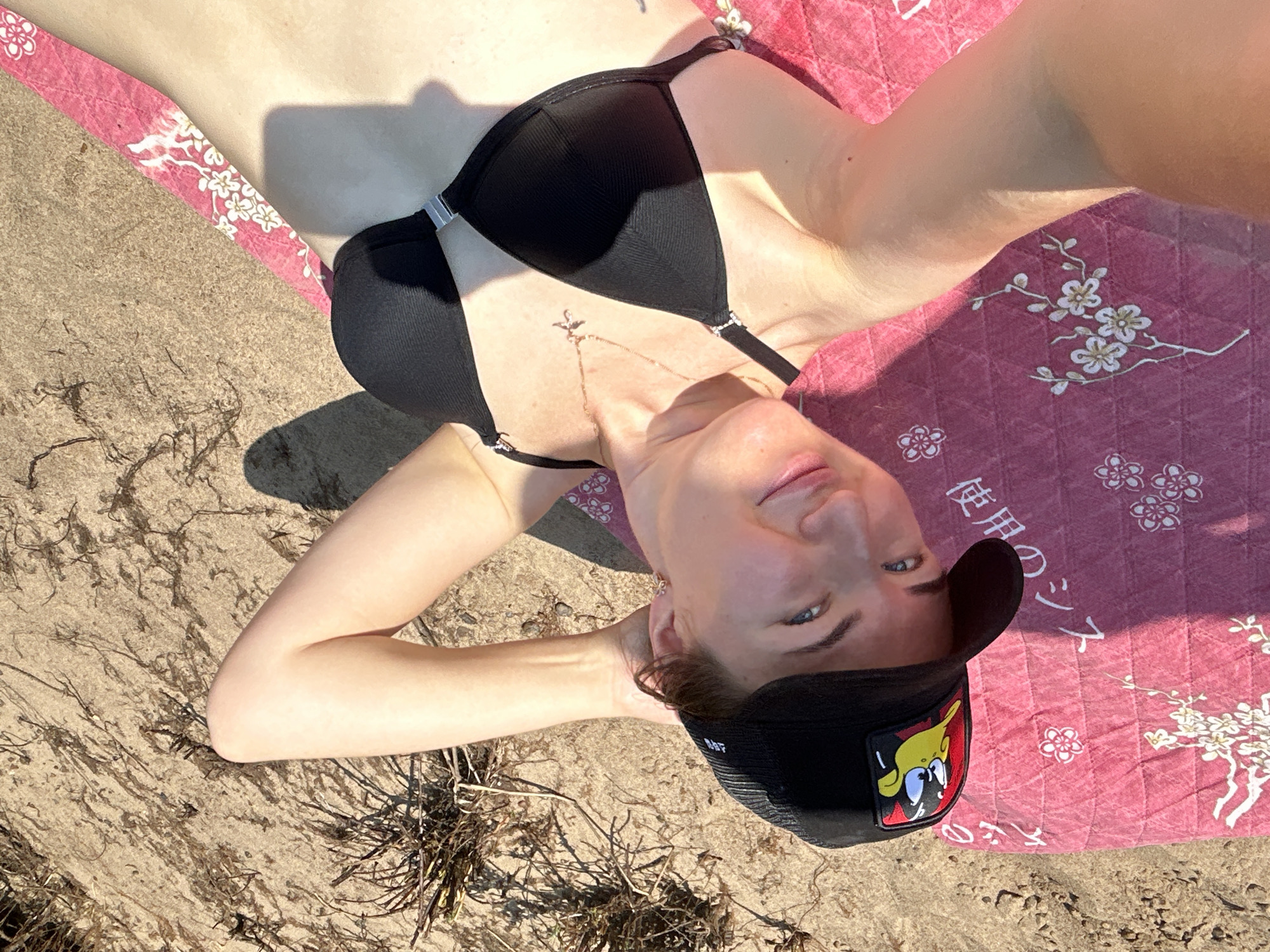 A woman lies on a pink blanket on a sandy beach. She is wearing a black bikini top and a black baseball cap with a cartoon duck logo. Her head is tilted back, and her left arm is resting on her head. She is looking towards the camera and smiling. The sand is dry and there are some dried plants and debris scattered across it. The lighting suggests it is a sunny day. The location is the city of Chaikovsky, Russia.