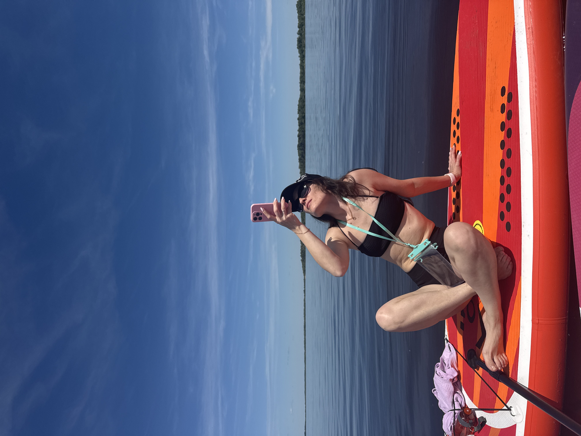 A woman is depicted kneeling on an orange and red paddleboard on a calm body of water under a bright, clear blue sky. She is positioned on the right side of the frame, facing left. The woman wears a black bikini top, dark shorts, a black cap, and sunglasses. She holds a pink smartphone in her right hand, appearing to be taking a photo or video. A light blue lanyard or strap is attached to her phone and hangs around her neck, and a clear, light blue-trimmed waterproof pouch is visible near her waist. Her left hand is resting on the paddleboard.
In the background, a narrow strip of green trees or land marks the horizon, separating the dark blue water from the lighter blue sky. The sky features some faint, wispy white clouds. Part of a second, purple or pink paddleboard is visible next to the main one. On the paddleboard near the woman's feet, a purple cloth or towel, a black paddle handle, and an orange-brown water bottle are secured. The scene suggests a sunny day activity on a large body of water, likely in Усть-Курдюм, Russia, consistent with the Volga River. No visible text is clearly discernible.