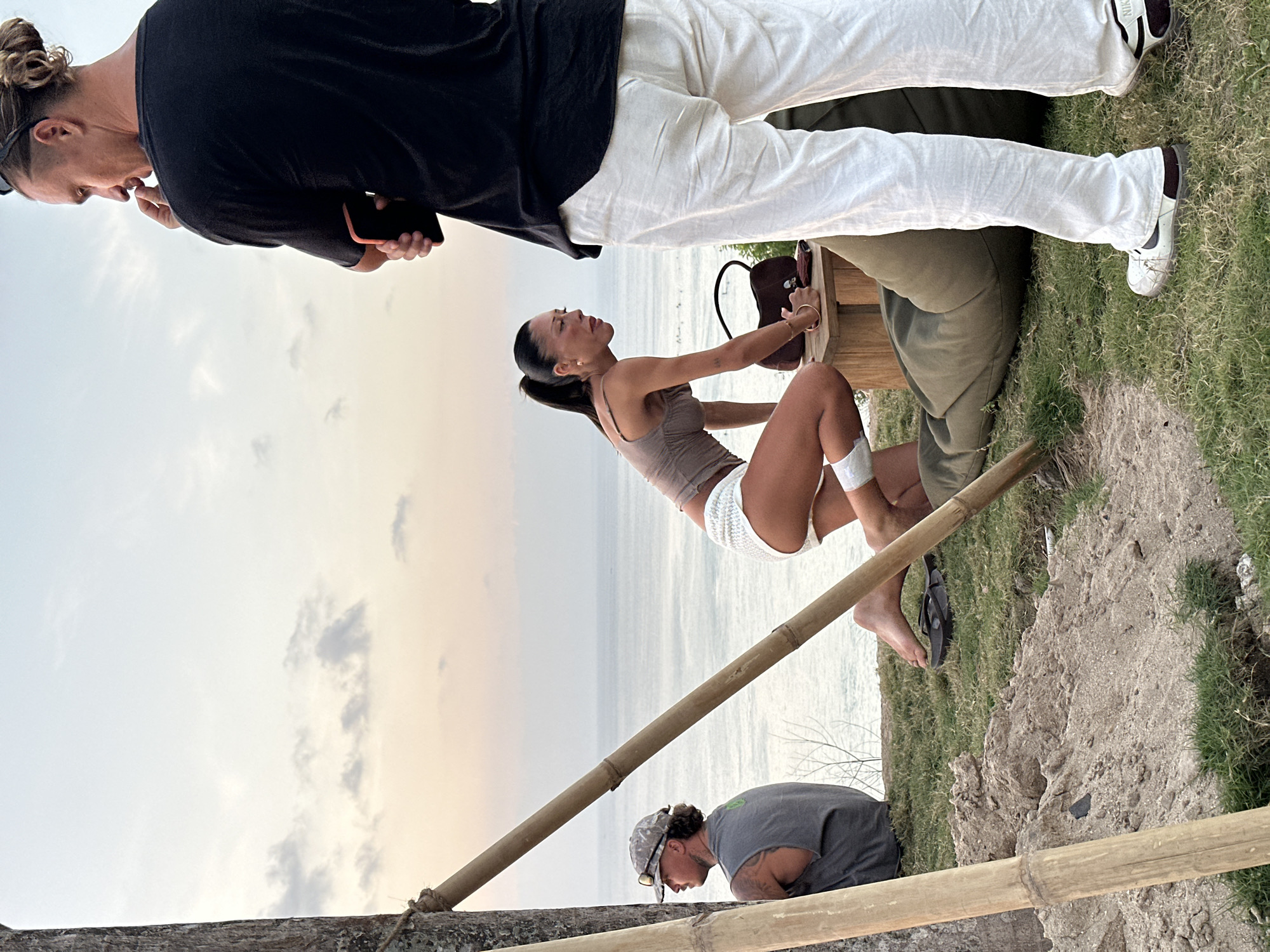 The media file shows a group of people near a body of water, likely the ocean. A man with his hair in a bun is holding a phone in his right hand. He is wearing a black long-sleeved shirt and white pants. Next to him, a woman in a taupe tank top and white shorts is sitting on what appears to be a wooden structure. She has a brown bag beside her. Behind the woman, another person is partially visible, wearing a green garment. In the foreground, a man wearing a grey t-shirt and a patterned cap is looking down. He has tattoos on his arms. The scene is set outdoors, with grass and a dirt path visible. The sky is overcast with hints of sunset colors. The overall mood appears casual and relaxed. There is no visible text in the image.