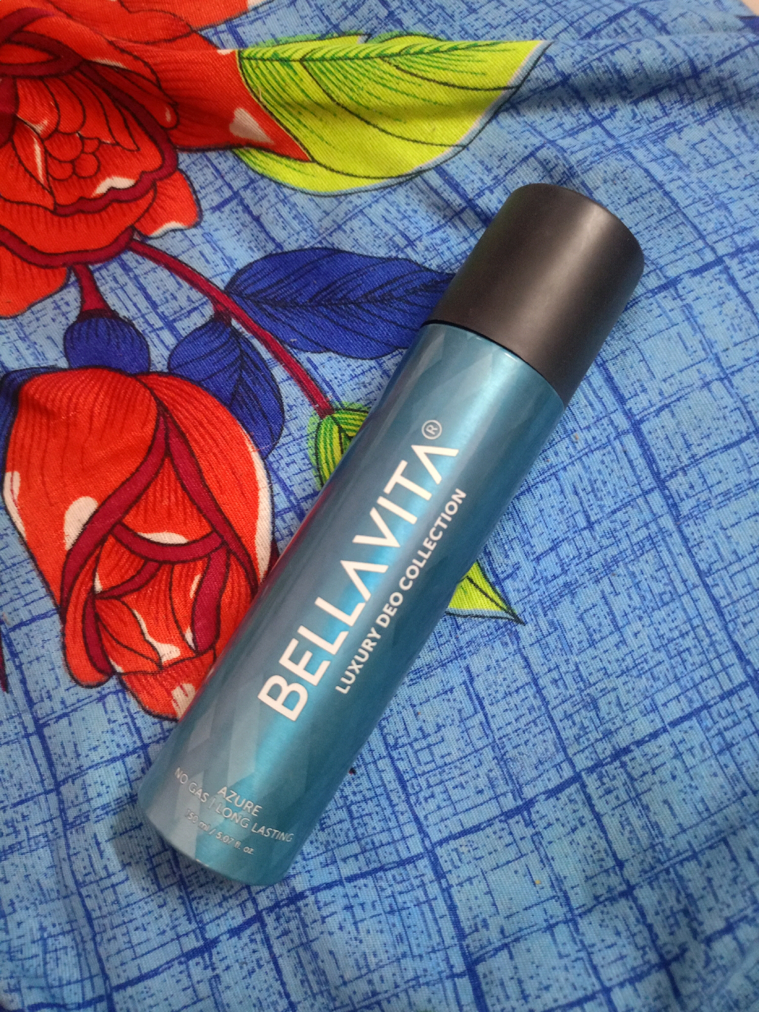 An overhead shot captures a teal-colored aerosol can with a black cap, resting diagonally on a blue fabric patterned with a red floral design. The can displays white text, reading "BELLAVITA LUXURY DEO COLLECTION" along its body, with smaller text below indicating "NO GAS | LONG LASTING", "AZURE", and "150 ml / 5.07 fl. oz.". The fabric itself is a vibrant blue with a stylized grid pattern overlaid with large, abstract red and green flowers, creating a busy and colorful backdrop. The lighting is even, suggesting an indoor setting, likely a bedroom or living area, typical of a residential environment. There are no people or animals visible in the frame. The composition is focused on the product, with the background fabric serving as a decorative element.