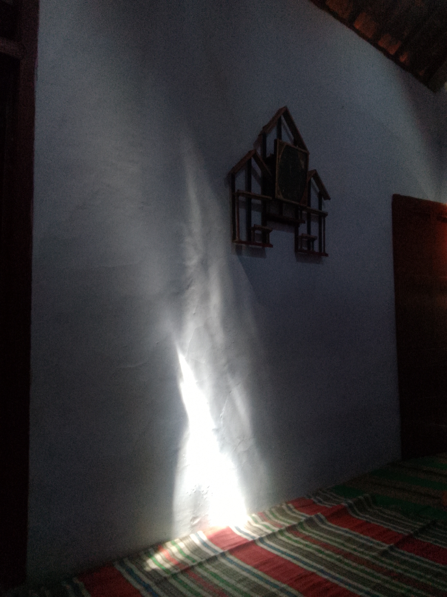 This image shows a close-up, indoor scene in Karangan, Indonesia. The focal point is a wall with a light beam illuminating it, creating a dramatic effect. On the wall, a wooden decorative clock in the shape of a house is mounted. Below the wall, a striped mattress or bedspread is visible, with red, green, and white stripes. The lighting is dim, with a strong shaft of light casting a bright, elongated shape on the textured wall. The overall impression is one of a humble, possibly rural, interior space.