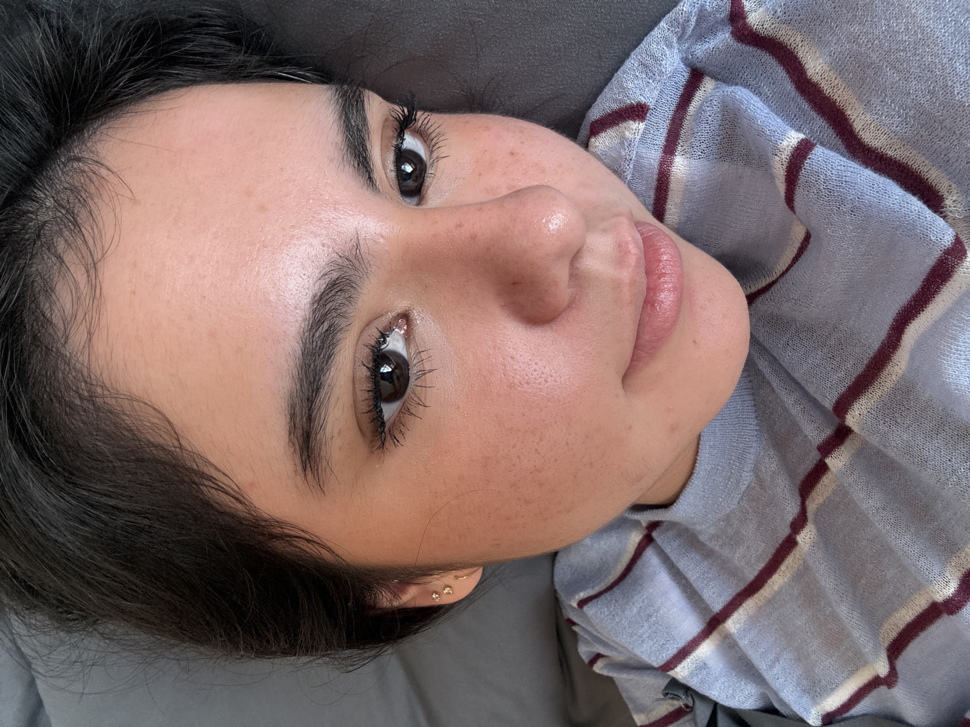 A close-up, angled photograph shows a young woman lying on a grey fabric surface. Her dark hair is visible around the top and left edges of the frame. She has fair skin with freckles, dark eyebrows, and long, dark eyelashes. Her eyes are brown. She is wearing small earrings. The woman is wearing a light blue and maroon striped shirt.
The location of the image is not apparent from the photograph and cannot be confirmed as San Jerónimo Chicahualco, Mexico.