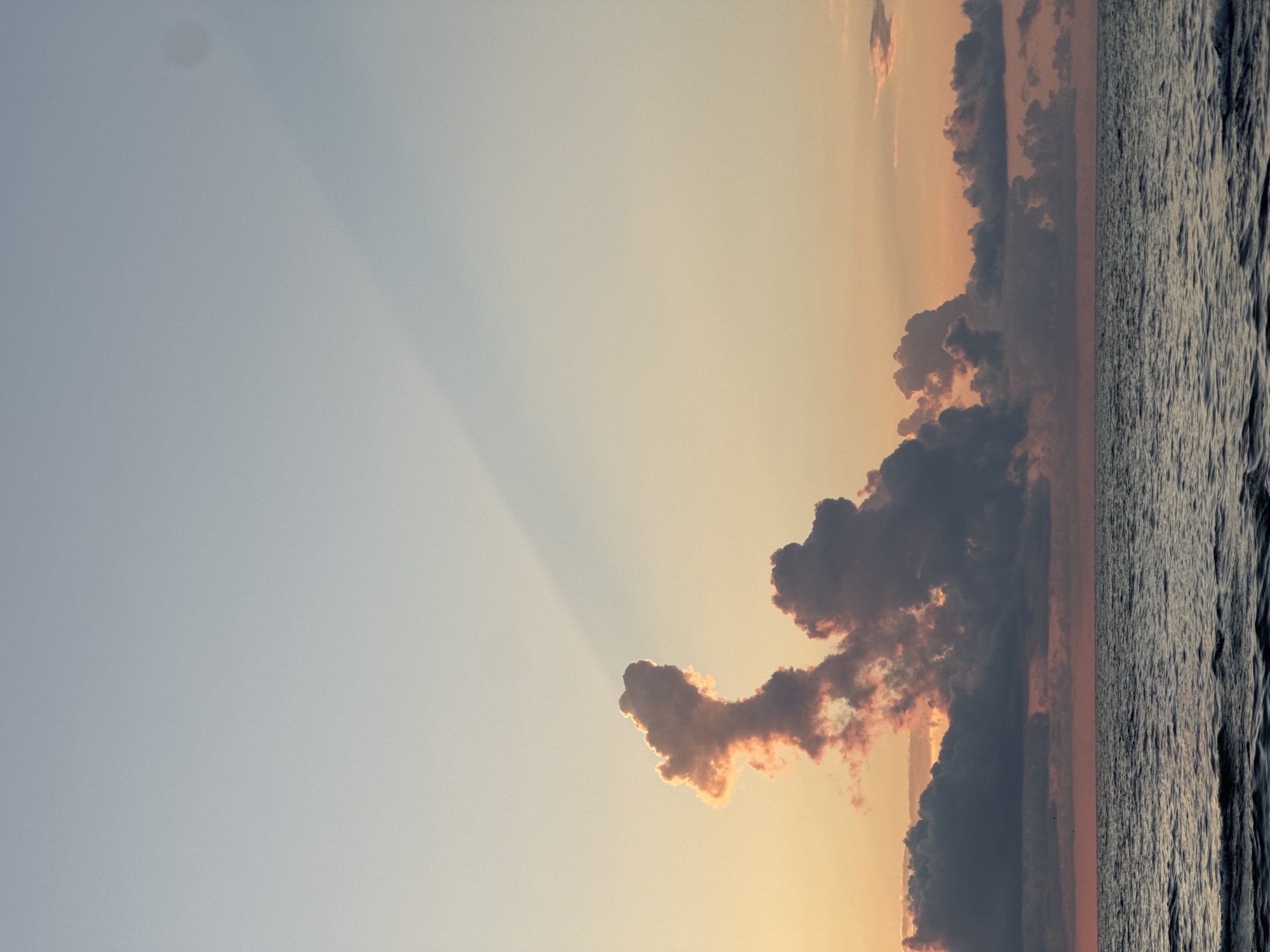 The image depicts a serene seascape at sunset, likely near Batumi, Georgia. The sky transitions from a pale blue at the top to warm oranges and pinks near the horizon, illuminated by the setting sun. A prominent, dark cloud formation sits in the middle ground, with one cloud in particular appearing to resemble a whimsical figure. A dramatic shaft of light pierces through the upper sky, cast from behind unseen clouds. The sea is calm, with gentle ripples reflecting the ambient light. No people or man-made structures are visible. The environment suggests a coastal location, characterized by the vast expanse of water and sky.