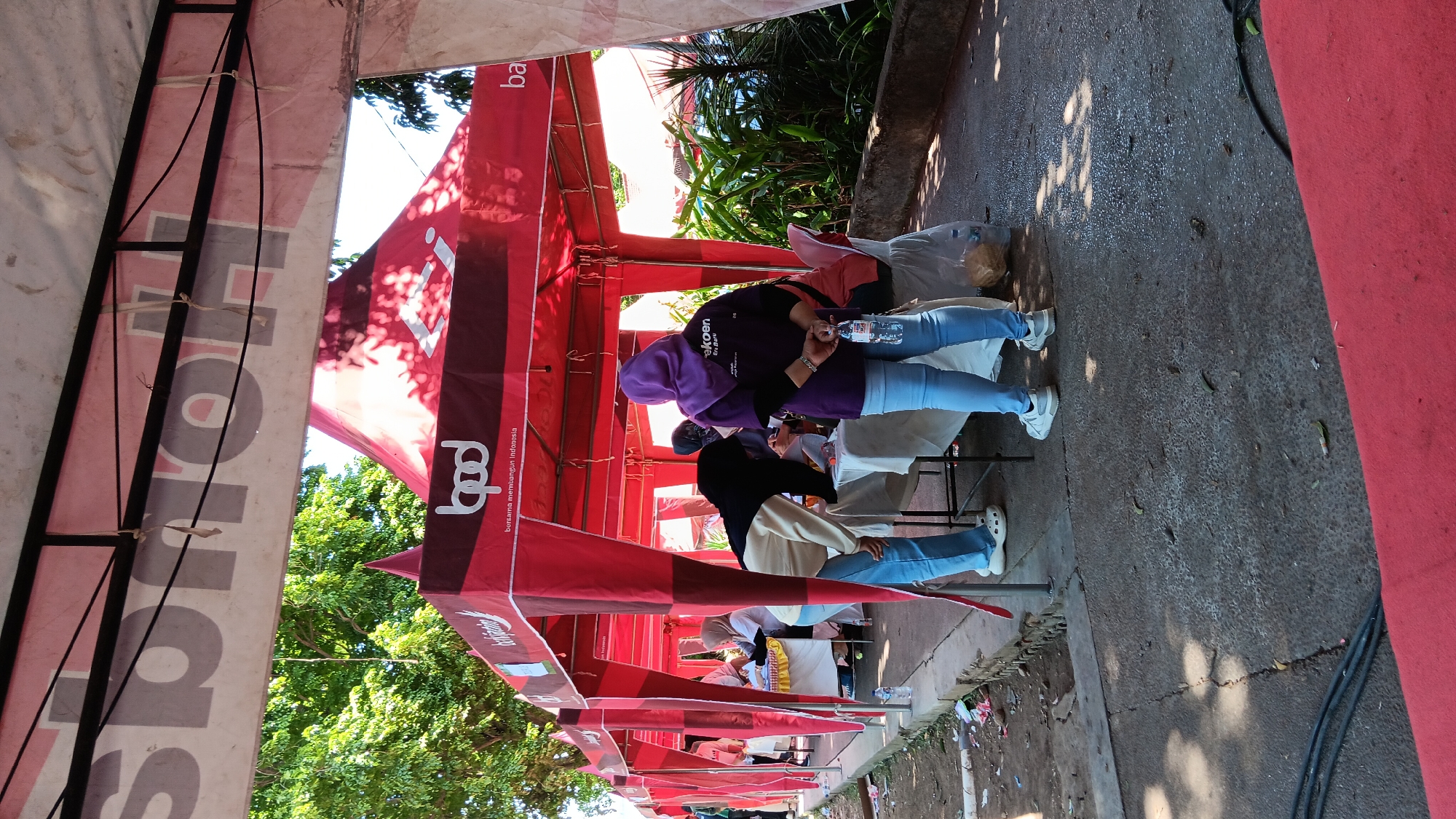 The image depicts an outdoor event with red tents set up, likely for a market or fair, in Nganjuk, Indonesia. In the foreground, a person wearing blue jeans and a purple hijab stands facing away from the camera, holding a water bottle and with their hands clasped behind their back. To their left, another person, also wearing blue jeans and a black and white jacket, is bent over a table. Several other individuals are visible behind tables under the tents. The ground is paved with concrete and there is some debris scattered around. Trees and foliage are visible in the background, suggesting a sunny day. The tents are branded with logos and text in Indonesian, one prominently featuring "bpd bersama membangun Indonesia."