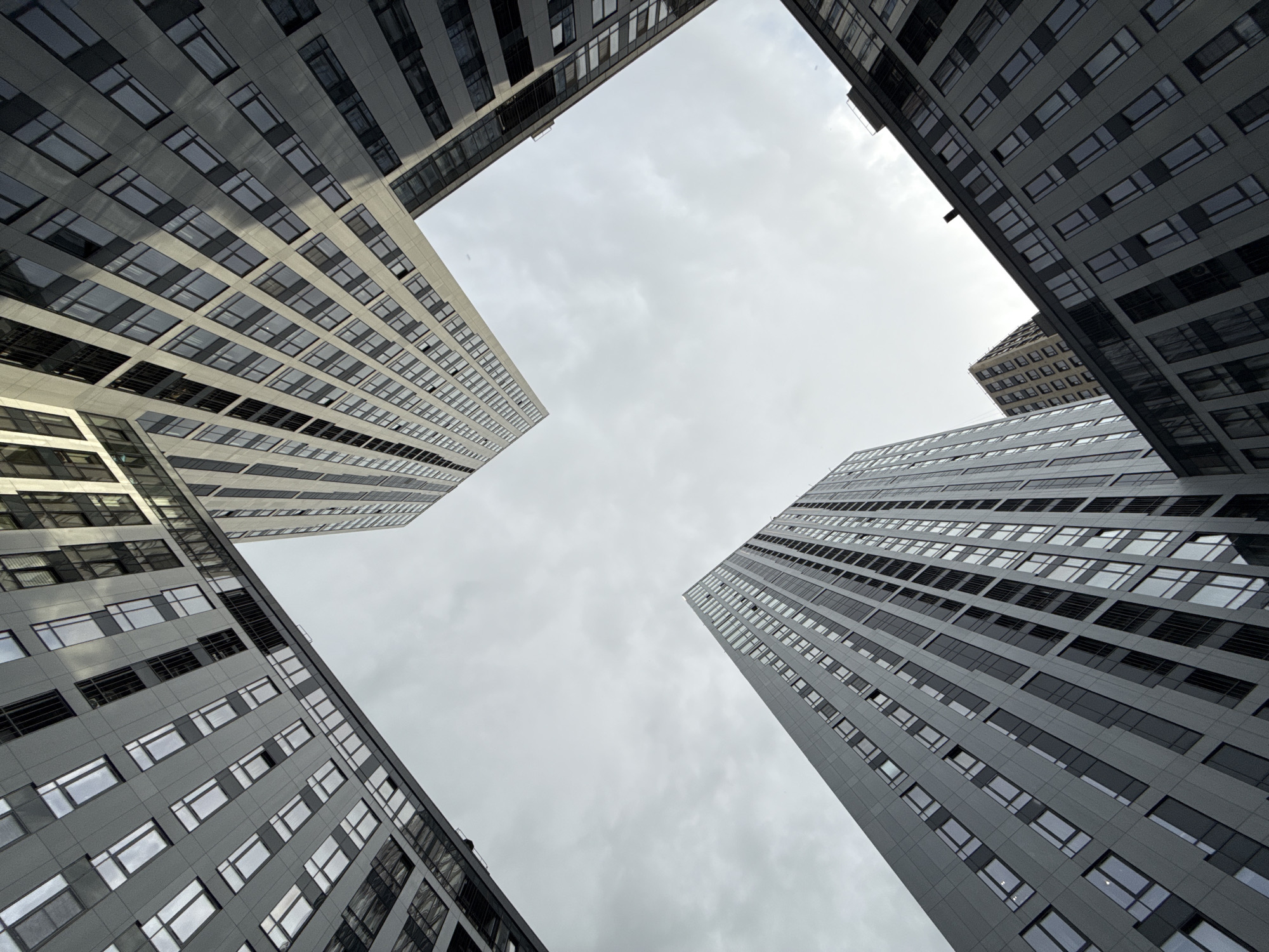 The image is a low-angle shot of several tall buildings in Moscow, Russia, arranged in a square formation. The buildings are modern, with a large number of windows visible. The sky is overcast. No people are visible.