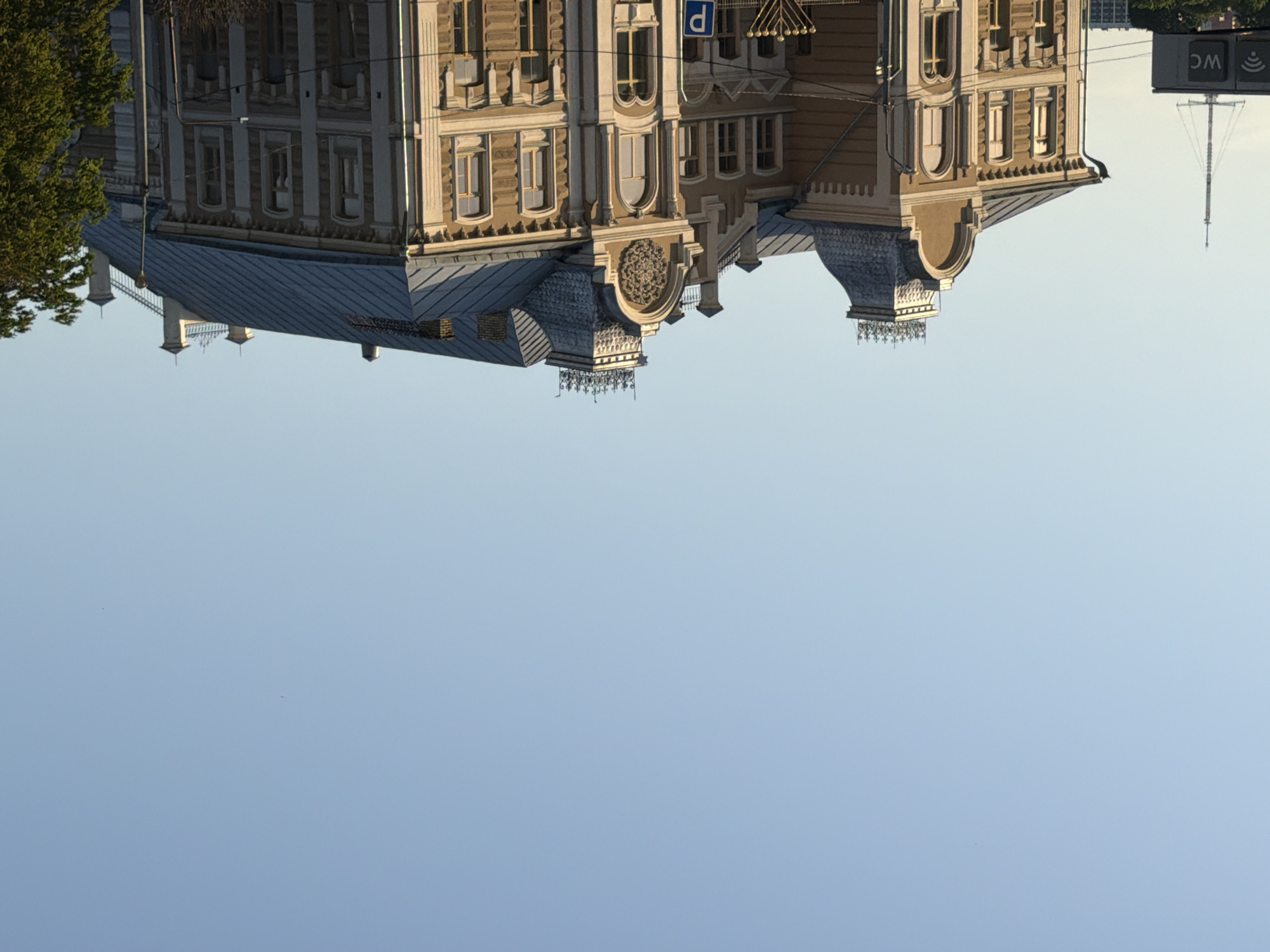 The media file displays an upside-down image of an ornate, historic building with a gray metal roof. The building's facade is a light brown or tan color, featuring multiple windows with decorative detailing, pilasters, and what appears to be a circular, ornate design on the upper section. The architecture suggests it is an older, possibly European structure. Adjacent to the building on the left is a large tree with green foliage. To the right of the building, a sign with the letters "WC" is visible, along with a symbol that resembles a Wi-Fi icon. Beyond the building and sign, a tall, slender tower or mast is visible, possibly a communication tower or flagpole. The sky is a clear, pale blue, suggesting daylight and fair weather. The orientation of the image implies it was captured with the camera inverted. No people are visible in the frame.