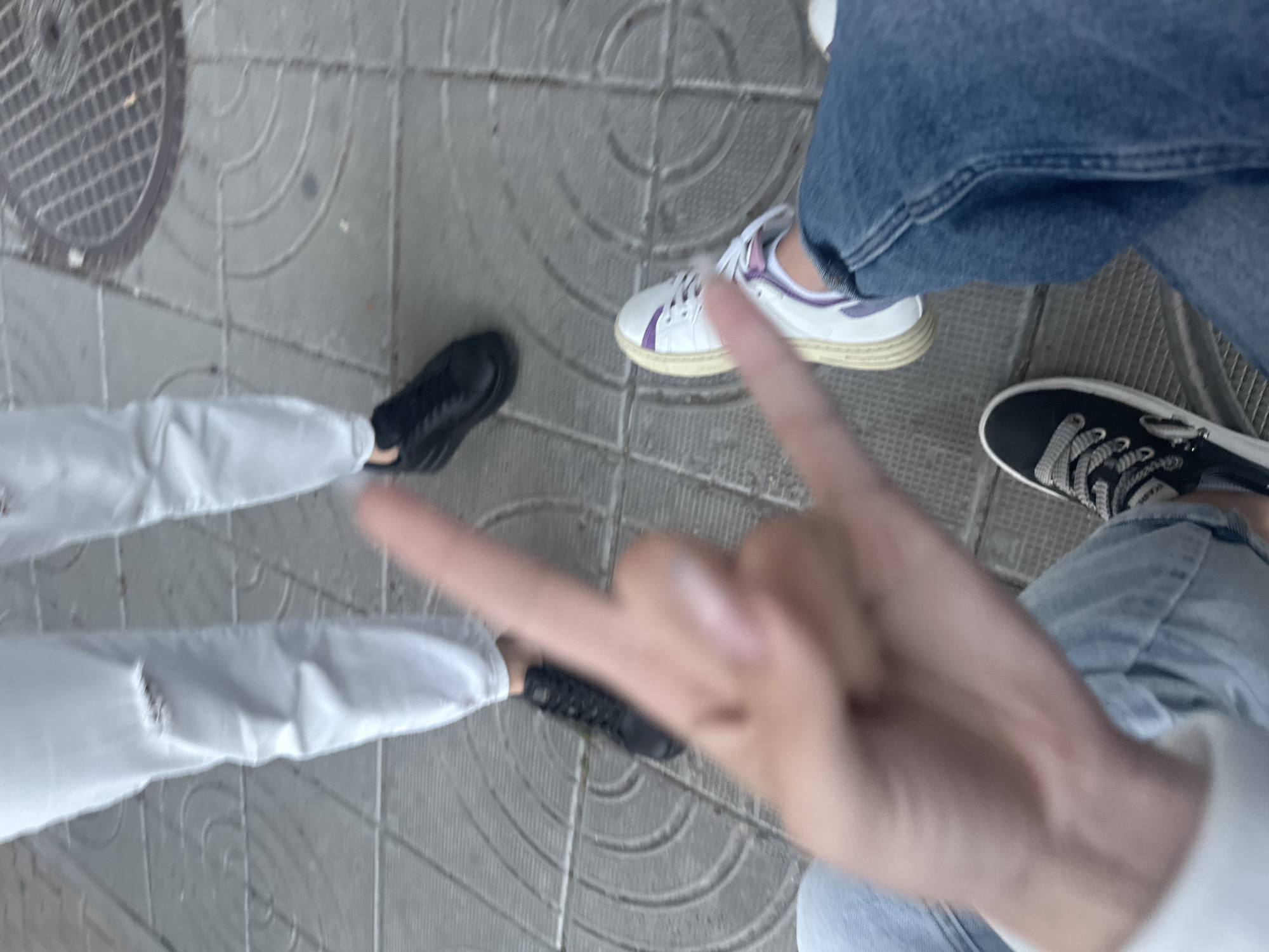 The image shows a close-up view of four people's legs and feet standing on a patterned cobblestone sidewalk in Shumen, Bulgaria. The people are wearing various types of shoes and pants. One person on the left is wearing ripped white jeans and black sneakers. Another person in the center is wearing white sneakers with purple accents and blue jeans. A third person, partially visible on the right, is wearing blue jeans and black sneakers with white laces. The foreground shows a hand making a "rock on" gesture. In the background, a circular metal grate is visible on the sidewalk. The lighting suggests it is daytime.