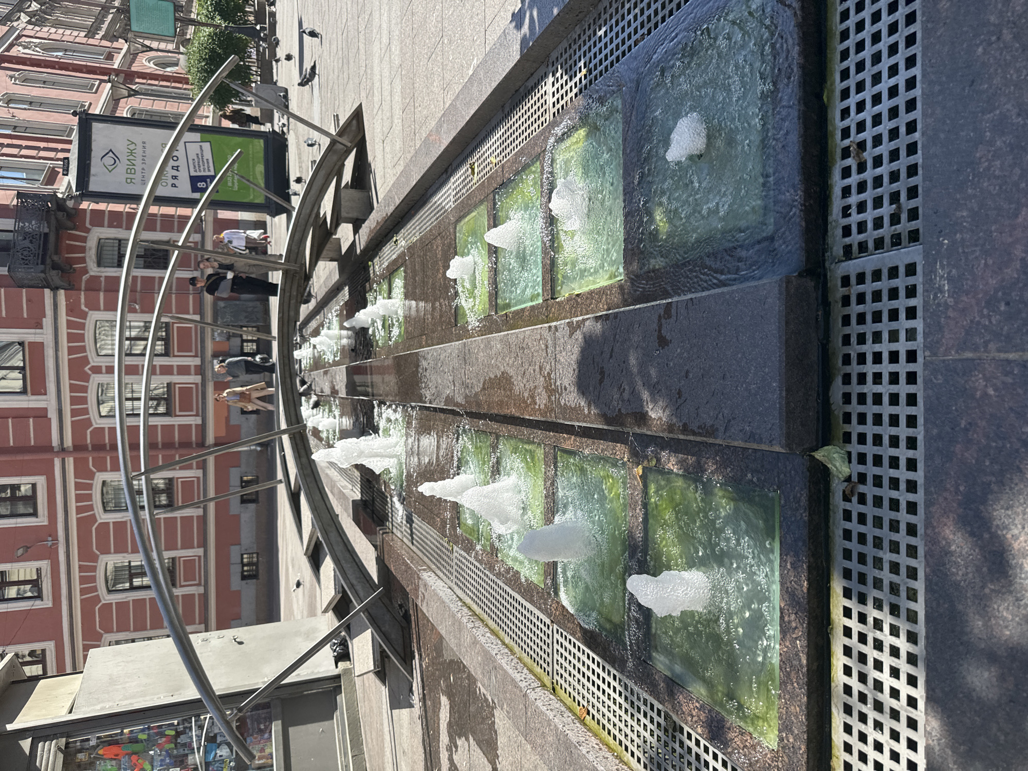 A sunny, outdoor scene in Saint Petersburg, Russia, depicts a decorative water fountain with multiple jets of water spraying upwards from square basins filled with green-tinted water. The fountain is set into a stone-paved area with metal grates. In the background, a multi-story building with red brick and arched windows stands along a city street. Several pigeons are scattered on the pavement, and a few people are visible walking in the distance. A large billboard with an eye logo and Russian text is also present. A curved metal structure arches over the fountain.